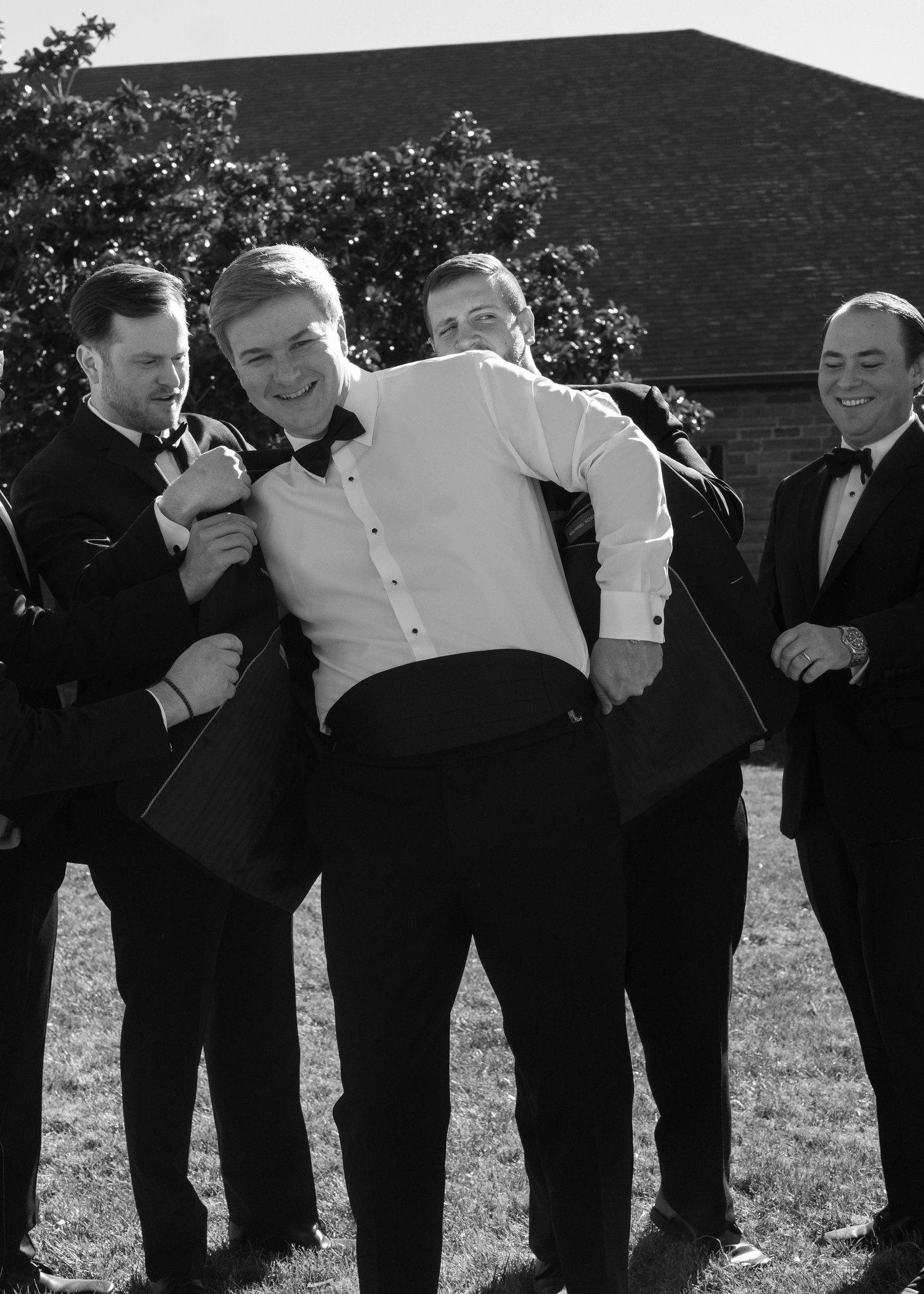 Groomsmen in black tuxedos posing together outdoors for a fun group photo.