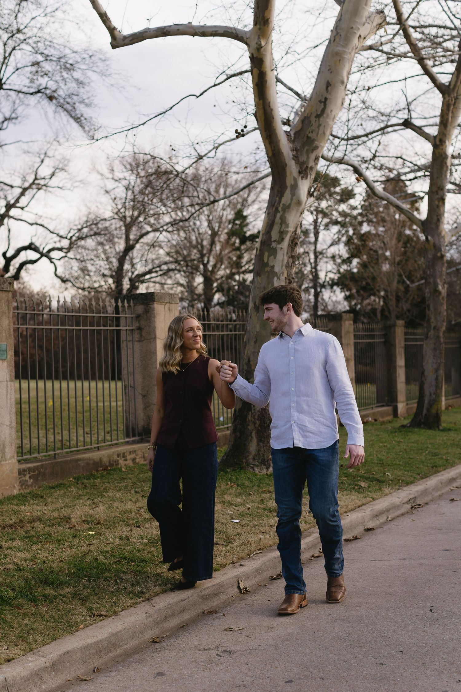 A couple walks hand in hand along a path near iron gates and bare winter trees.