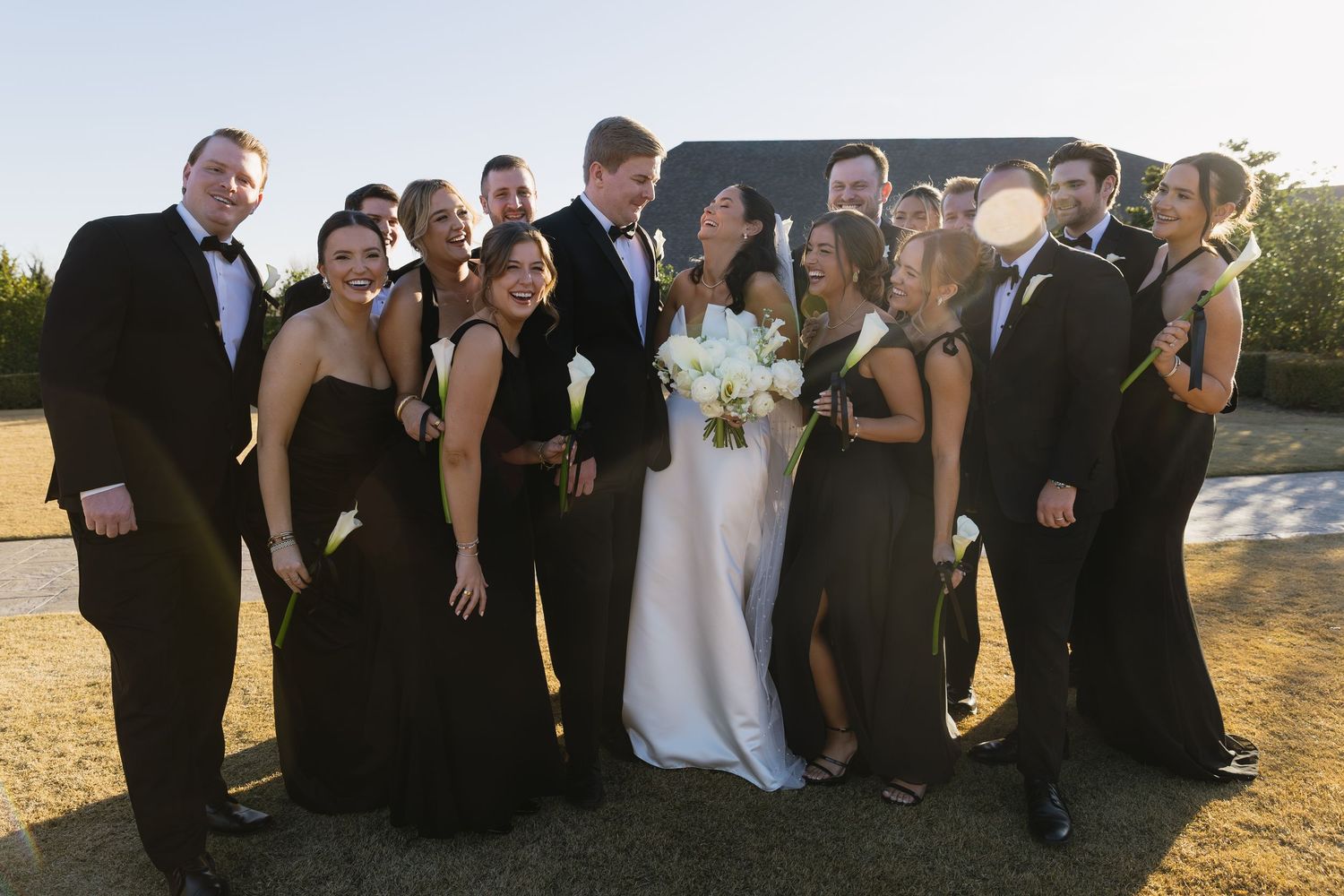 A wedding party in formal black attire and white gowns poses together outdoors at sunset.