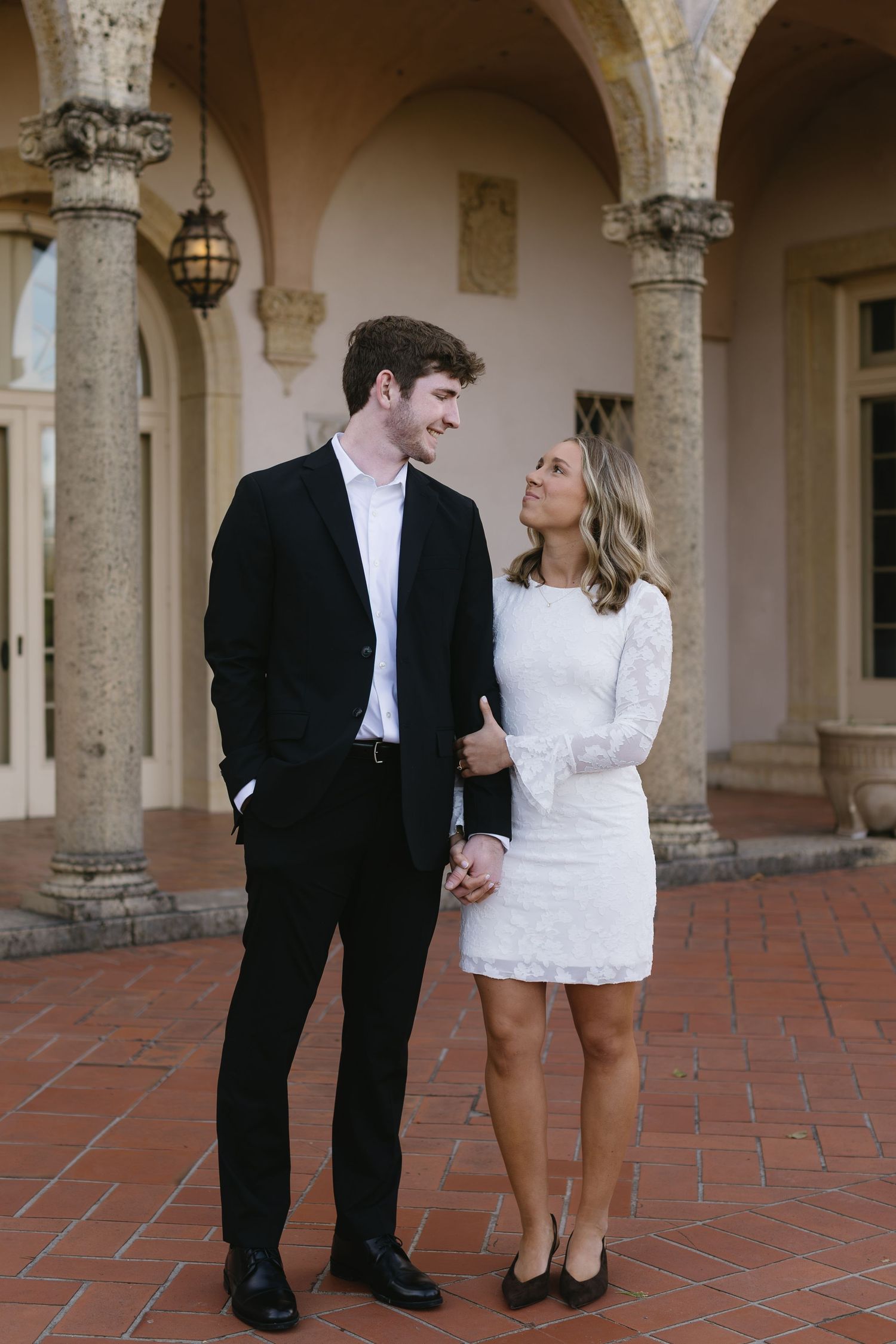 A romantic couple in formal attire share a tender moment under an arched colonnade with terracotta flooring.