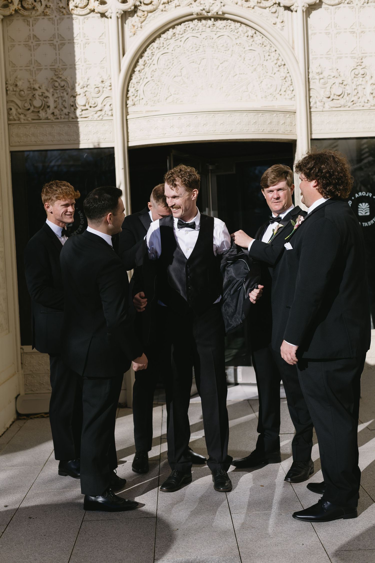Groomsmen in black tuxedos gathered in an ornate hallway.