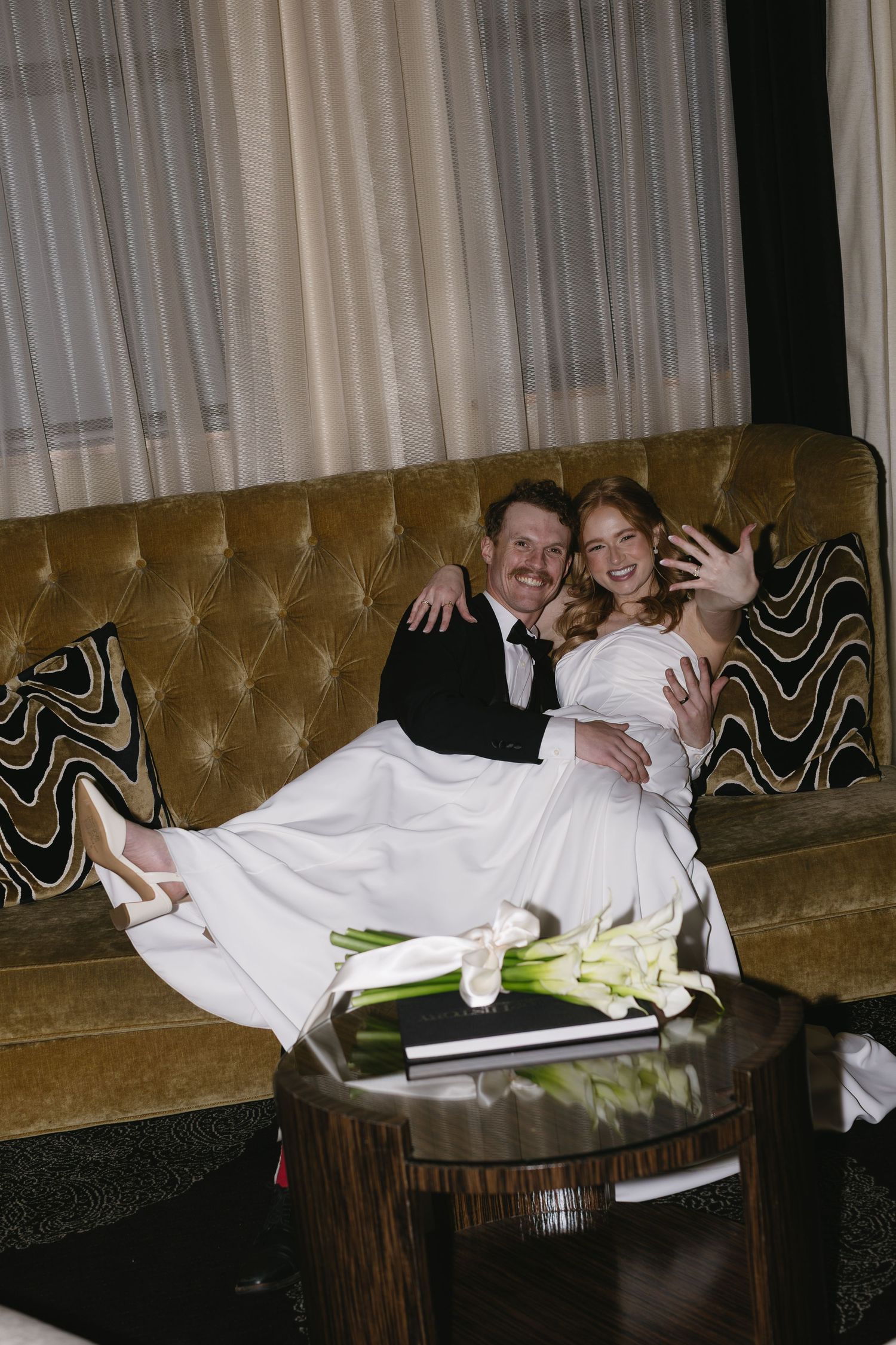 Wedding couple relaxing on vintage brown leather couch with calla lily bouquet.