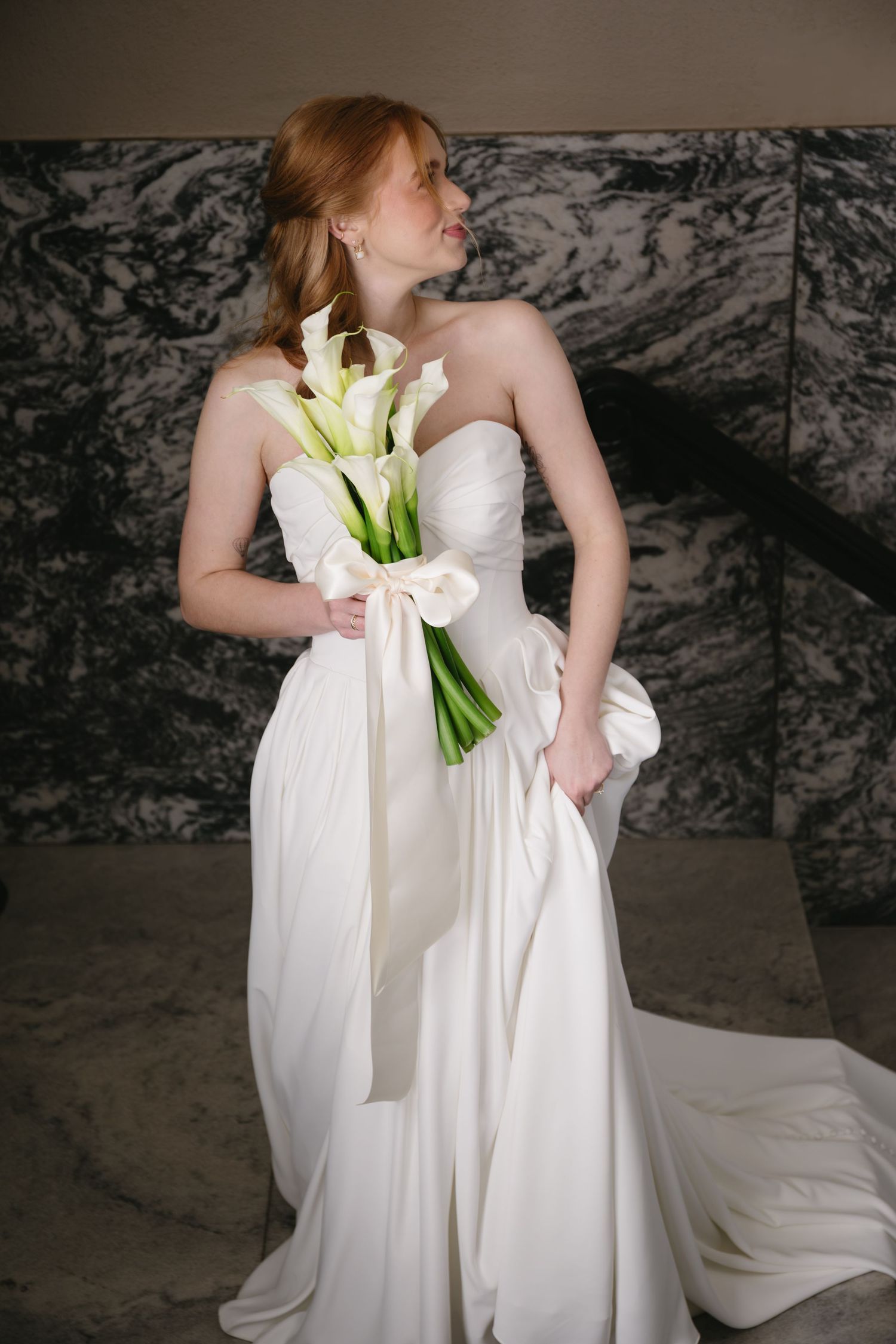 Elegant white wedding dress with calla lily bouquet against dark textured background.