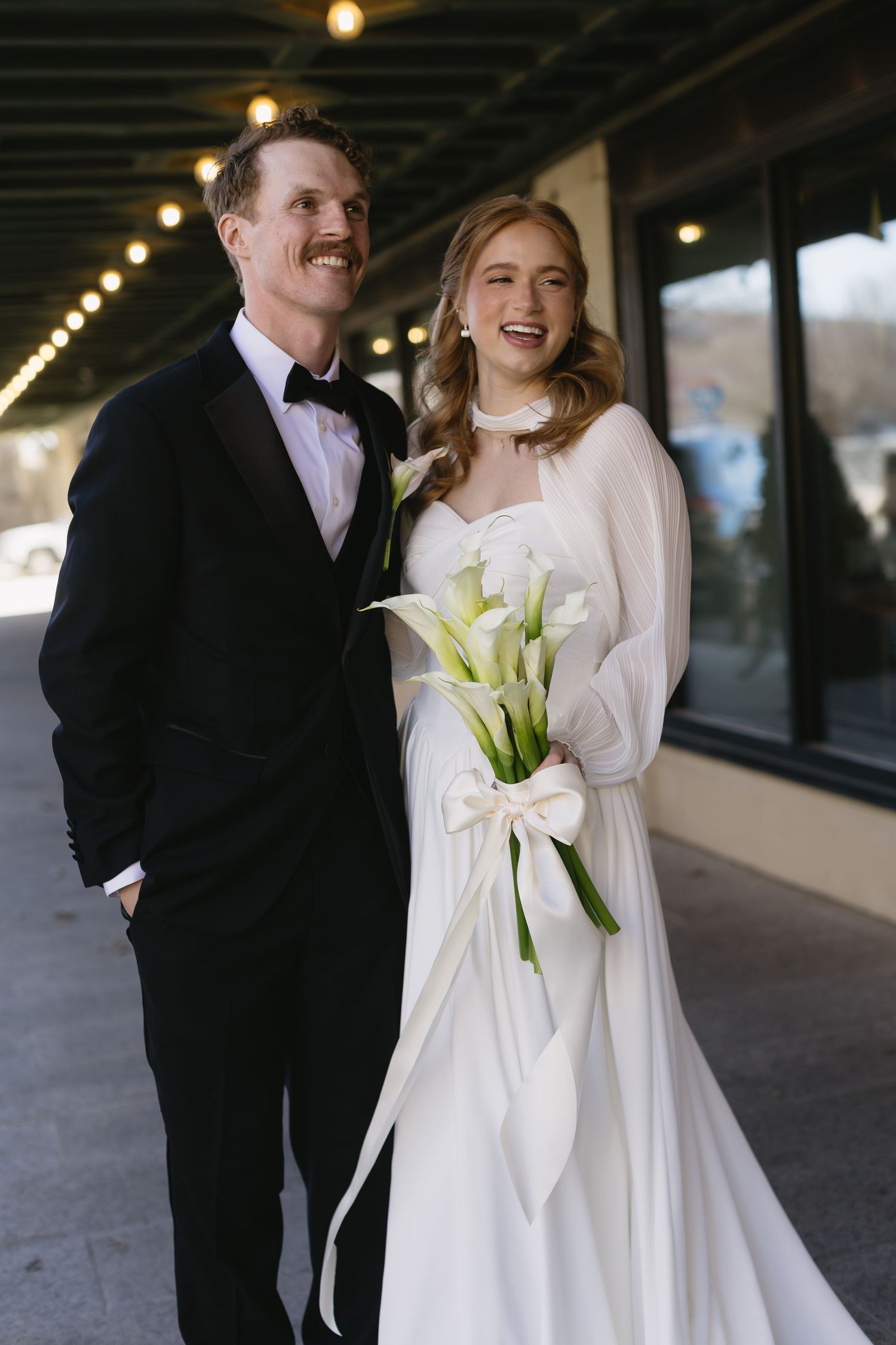 Newlyweds sharing romantic moments in outdoor covered walkway with calla lily bouquet.