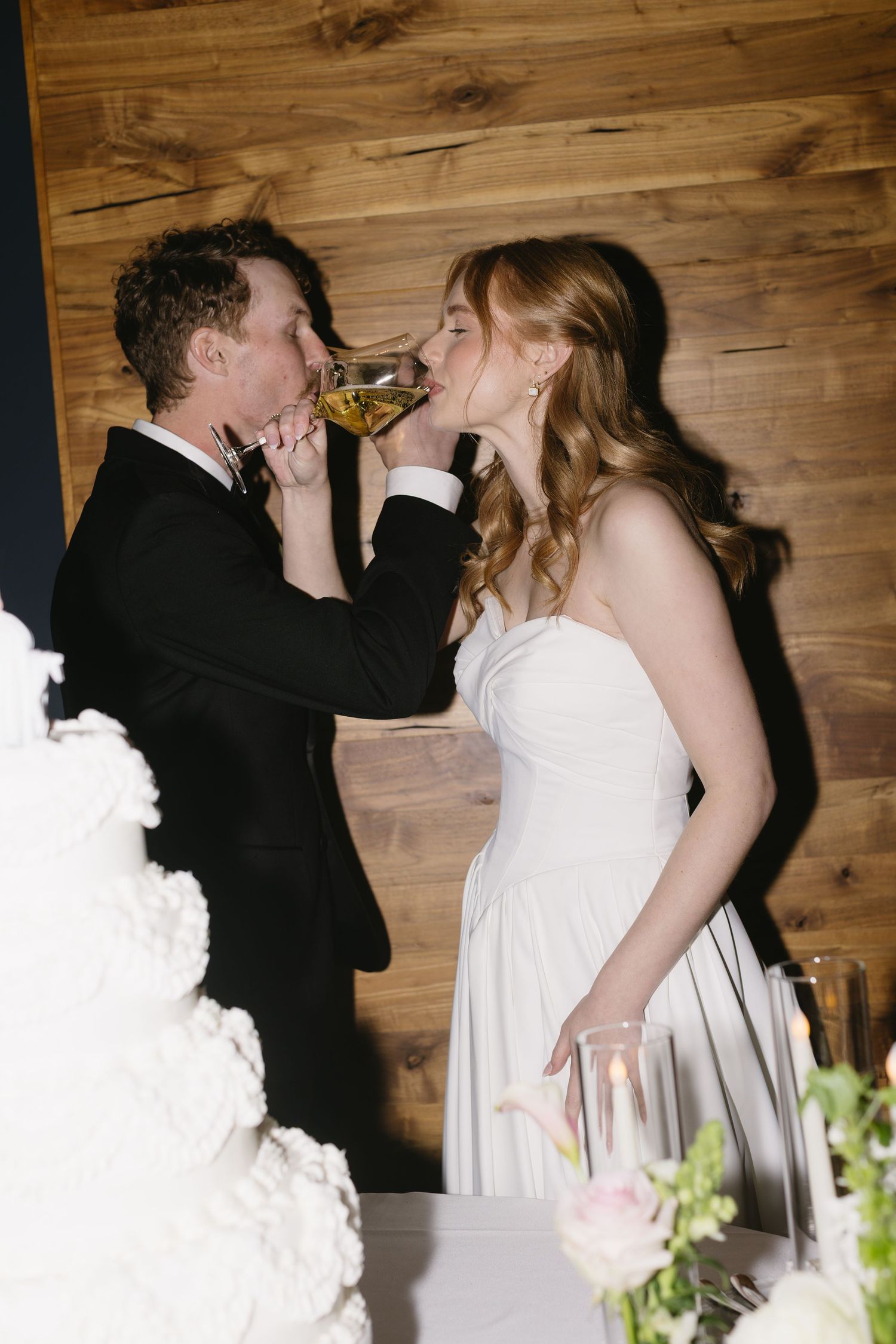 A couple shares a romantic champagne toast at their wedding reception against a rustic wooden wall backdrop.