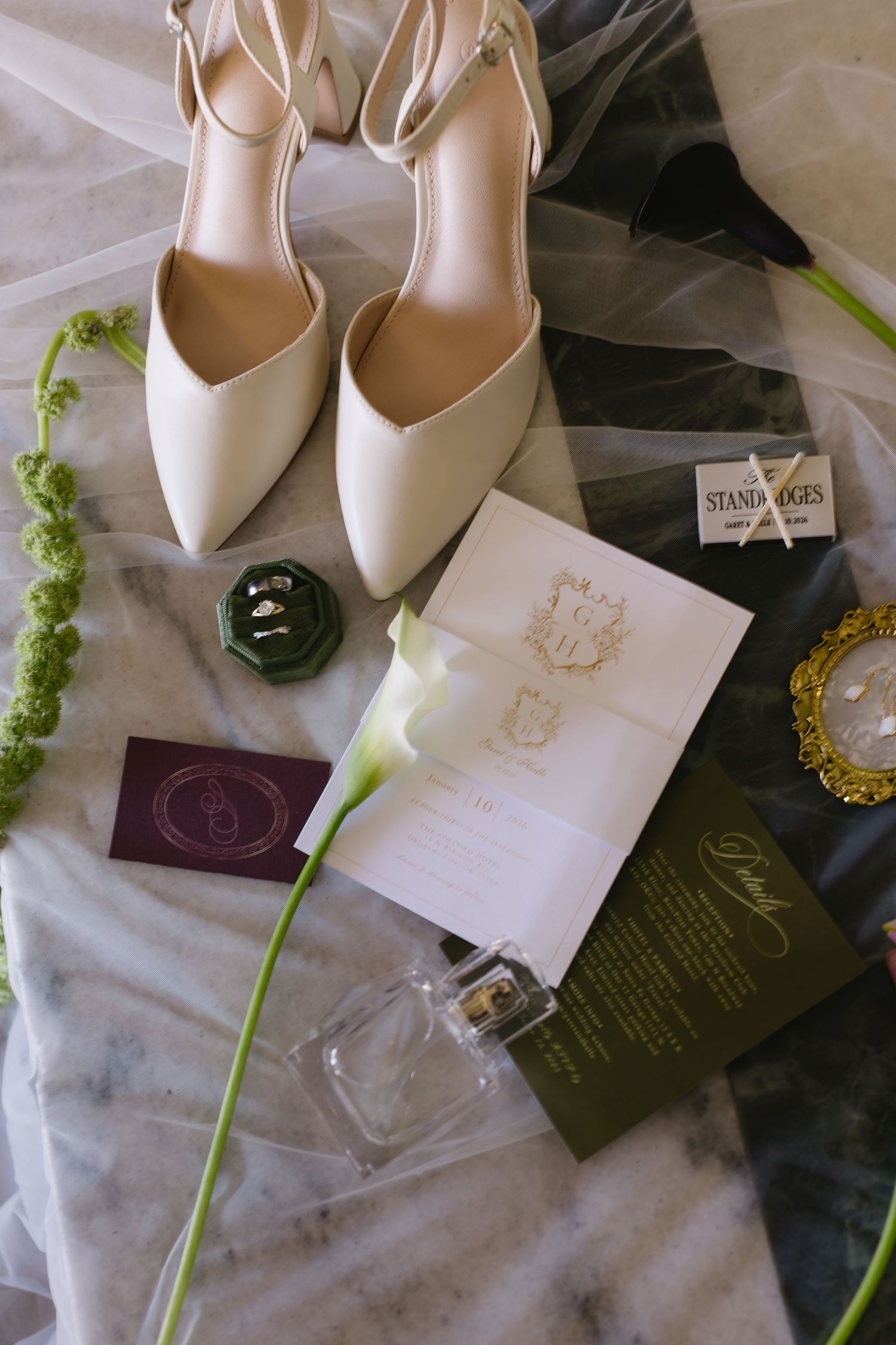 Wedding details flatlay with white heels, invitation, perfume bottle and calla lily on tulle fabric.