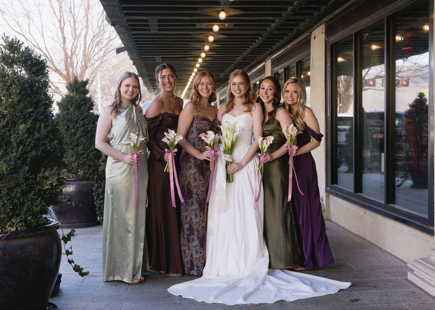 Bridesmaids in purple and brown dresses pose together with the bride in a white wedding gown outdoors.
