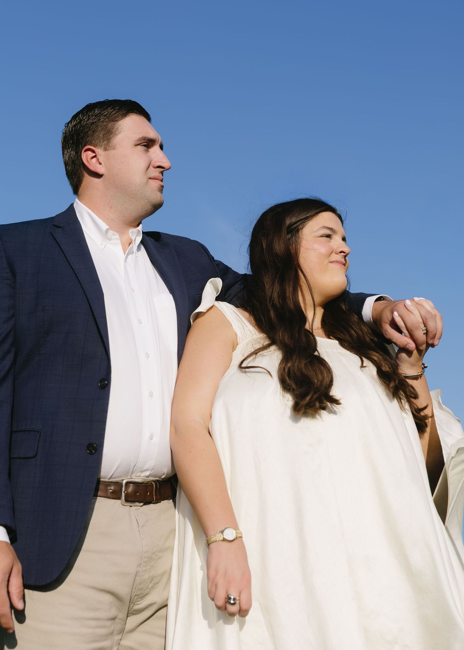 Elegant couple in formal wear posing against bright blue sky during golden hour.