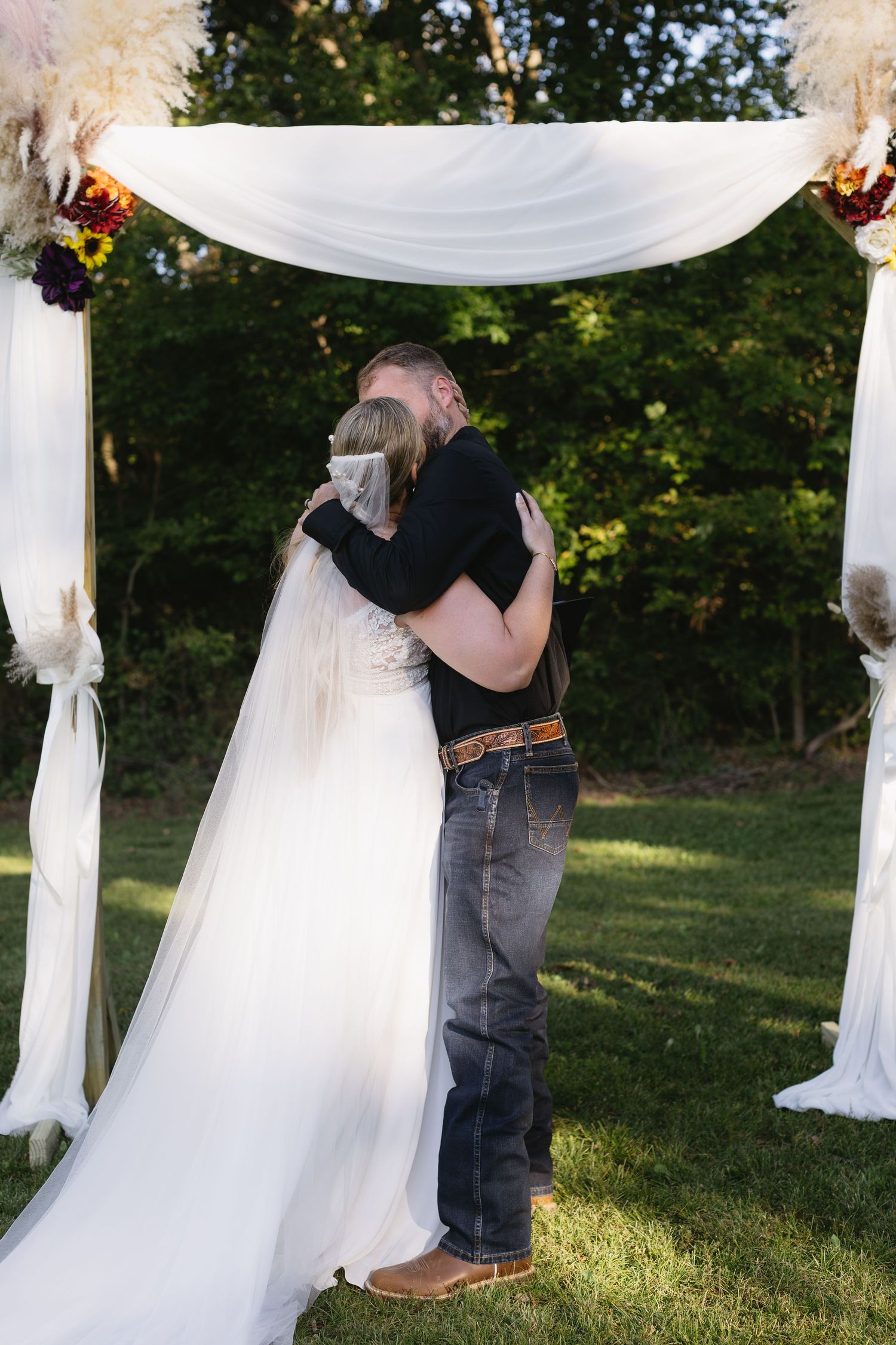 Wedding ceremony under a white draped arch with guests gathered outdoors.