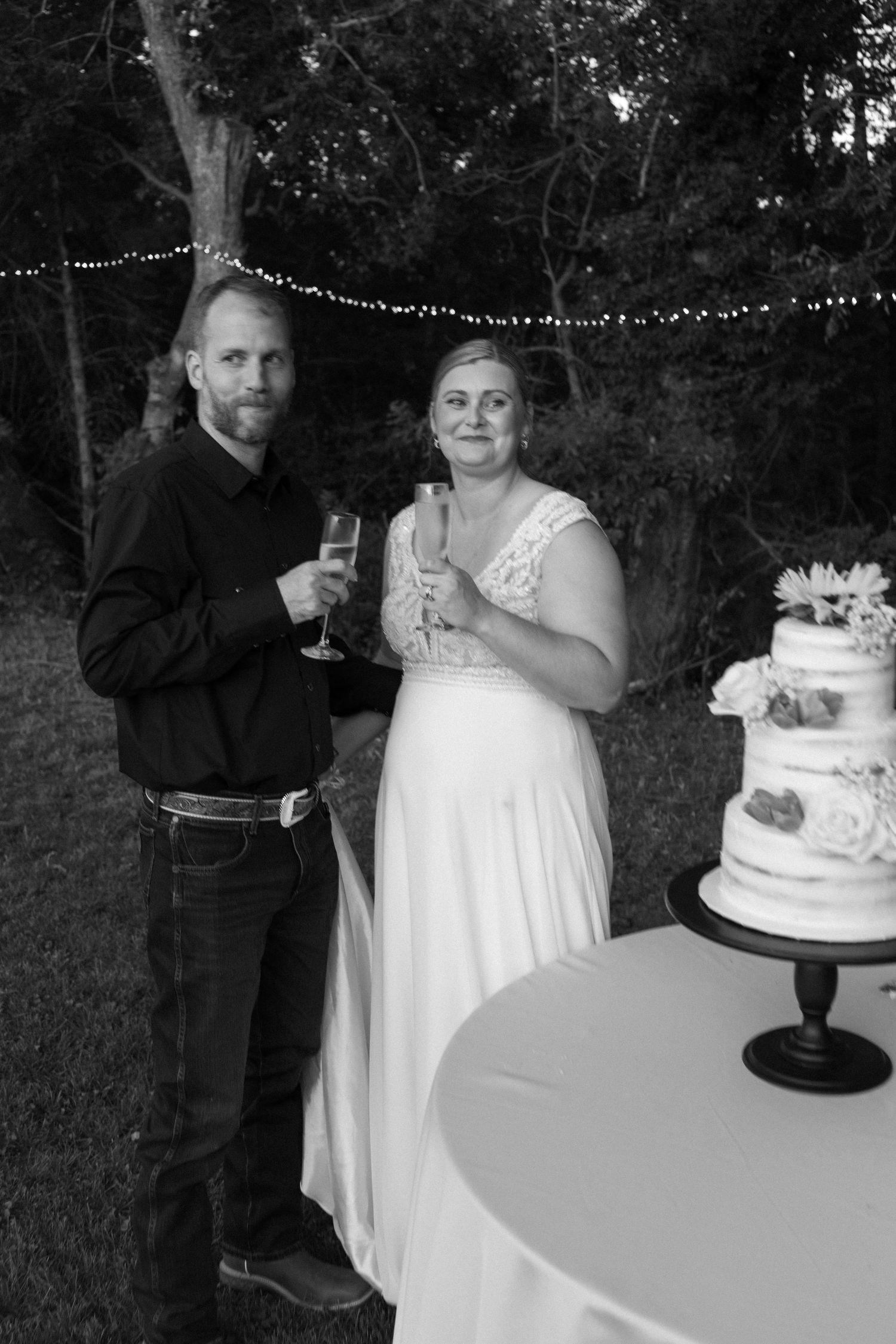A black and white photo of newlyweds toasting by their wedding cake under string lights.