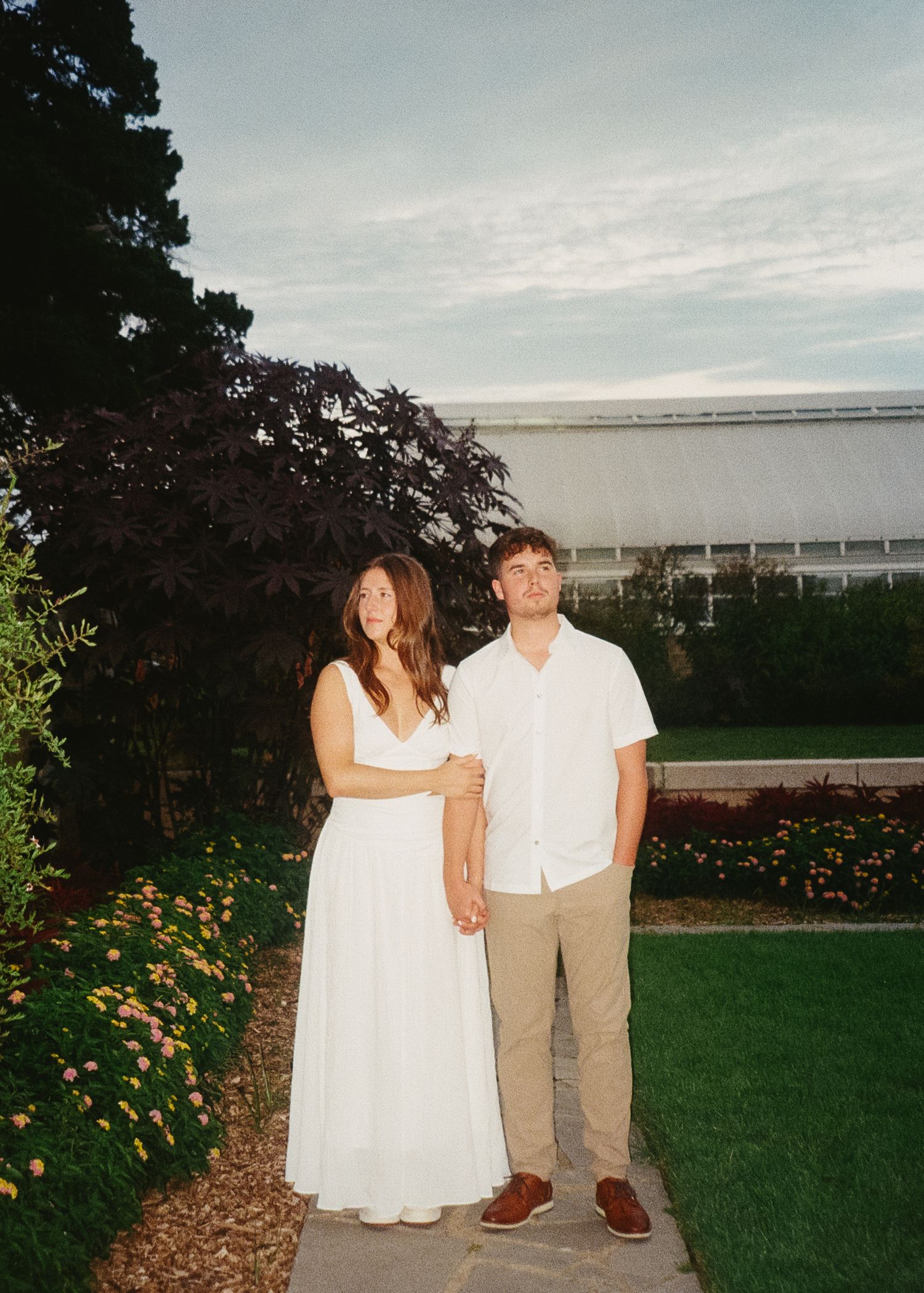 A couple stands together on a grassy lawn with flowers and buildings in the background at dusk.
