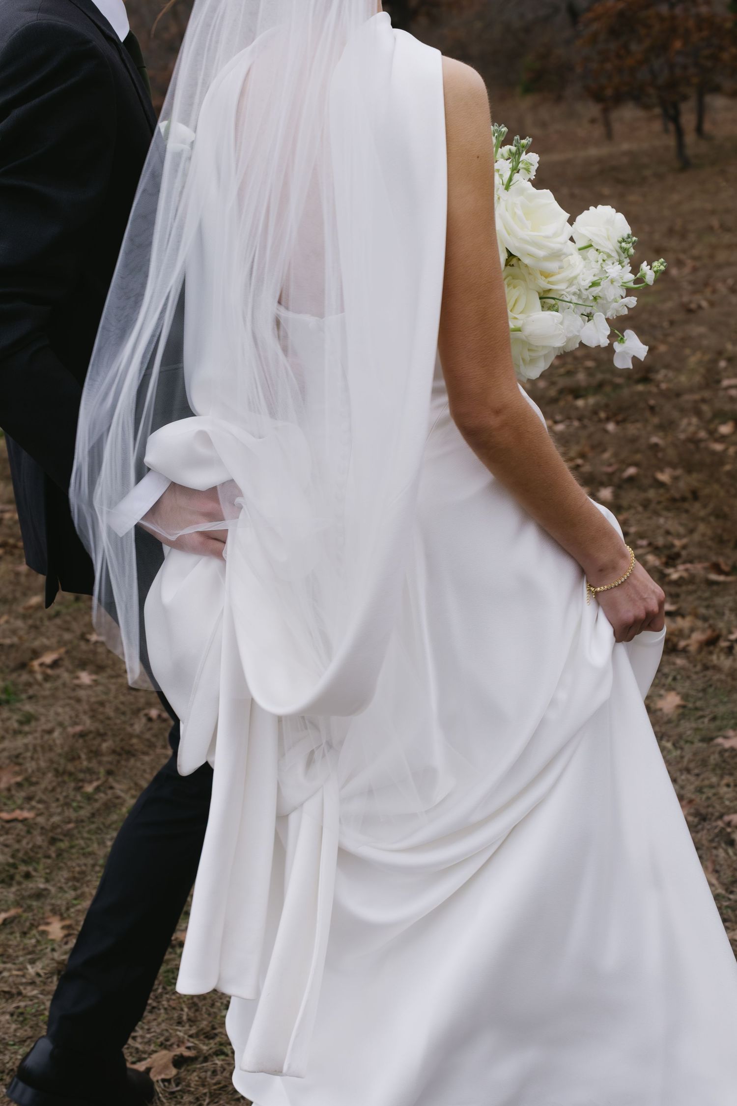 Close-up detail of bride's flowing white dress and veil as couple walks together outdoors.