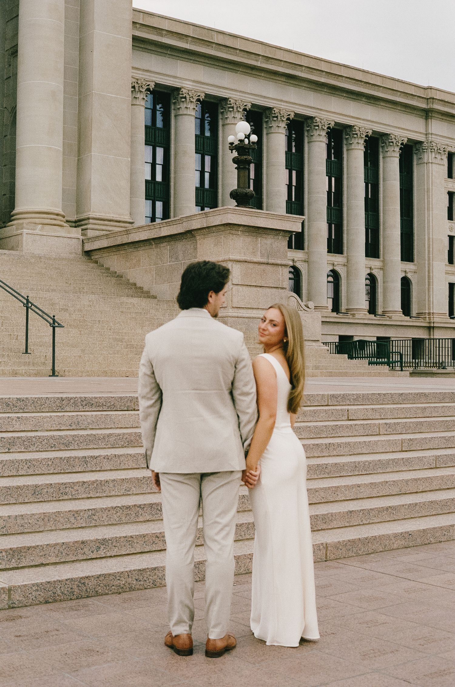 A couple in formal wear stands on grand stone steps of a classical building.
