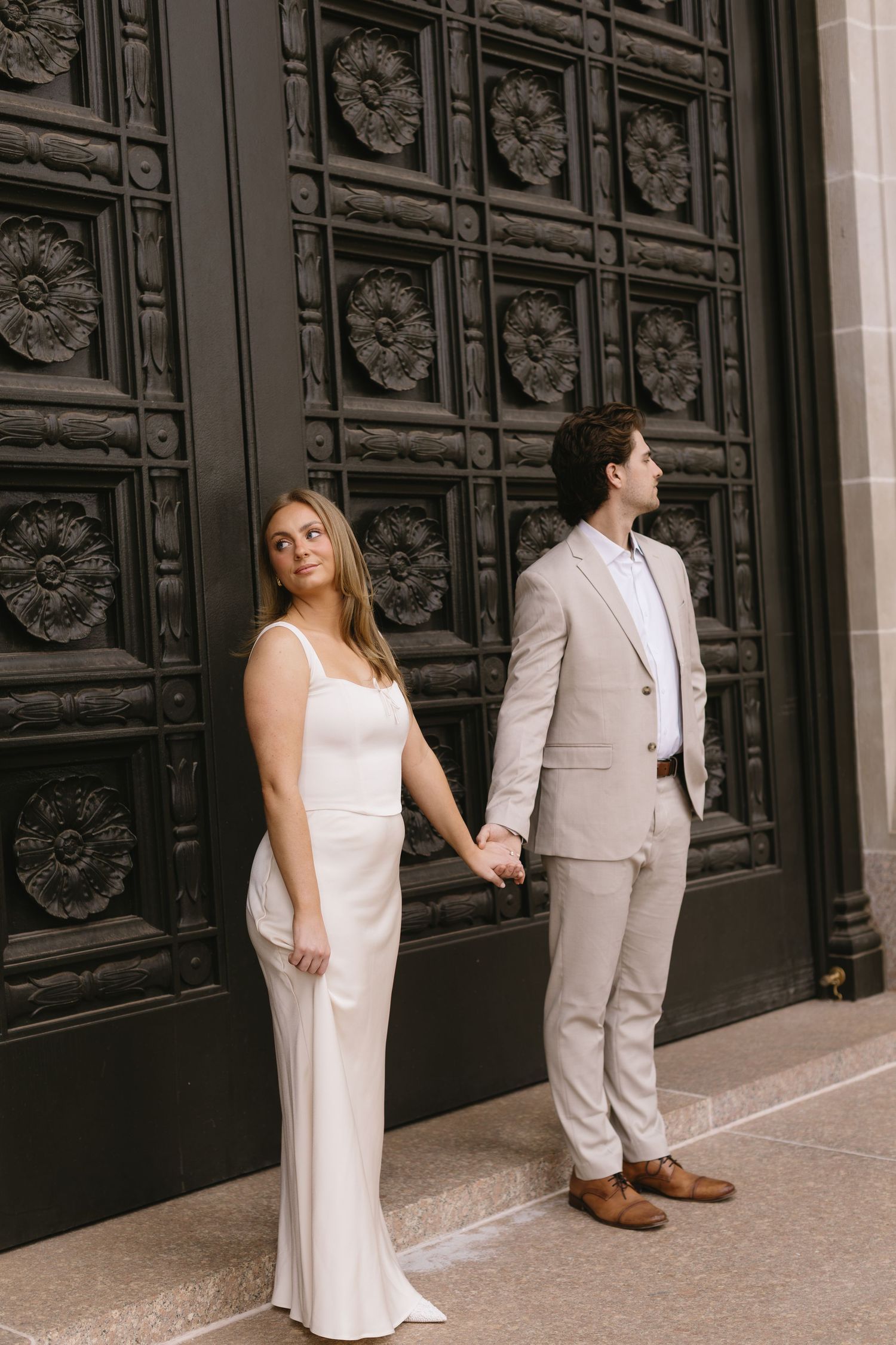 A couple in wedding attire poses against ornate black doors during an elegant photoshoot.