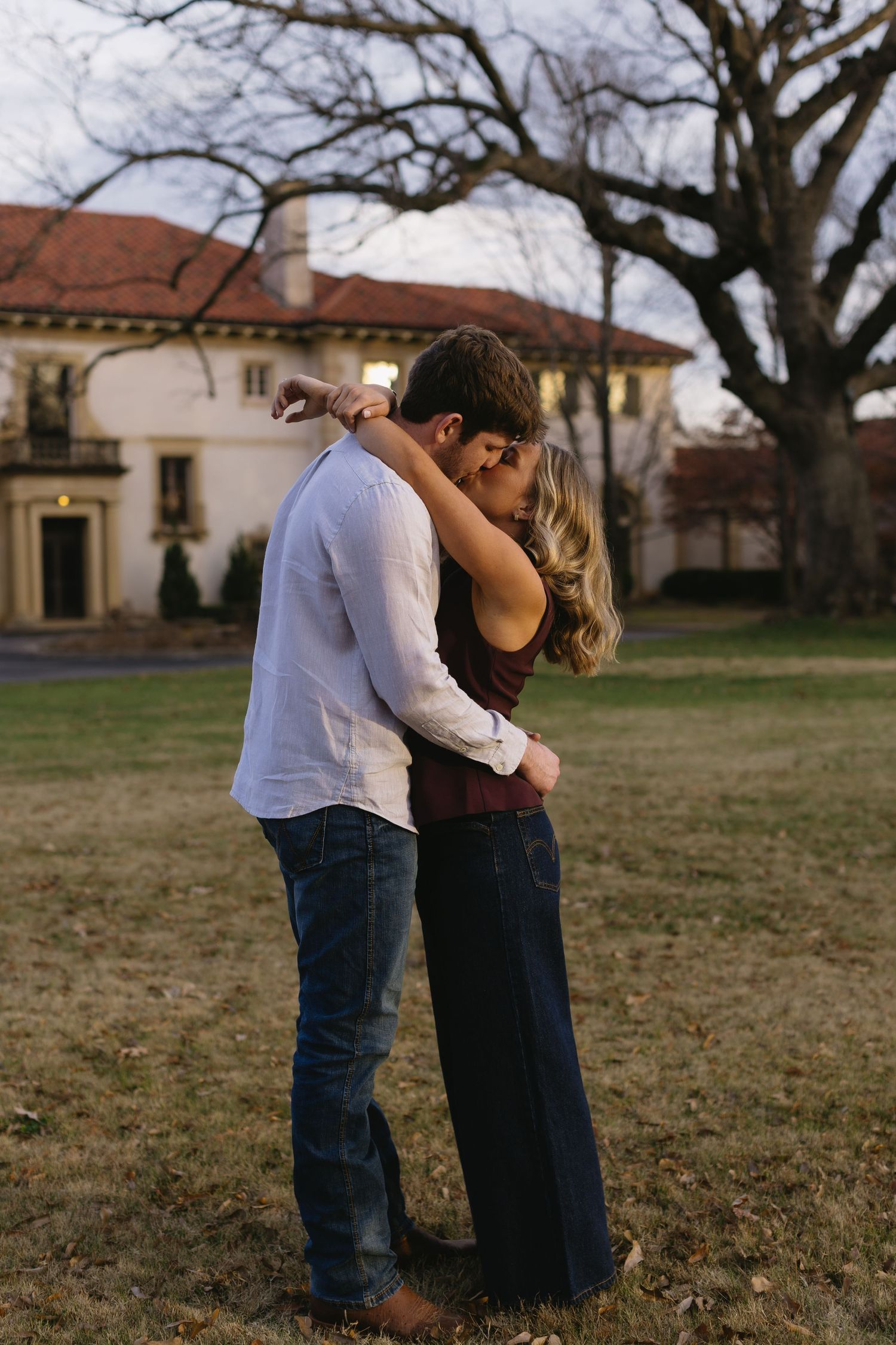 Romantic couple embracing in front of Spanish-style buildings and bare trees during golden hour.