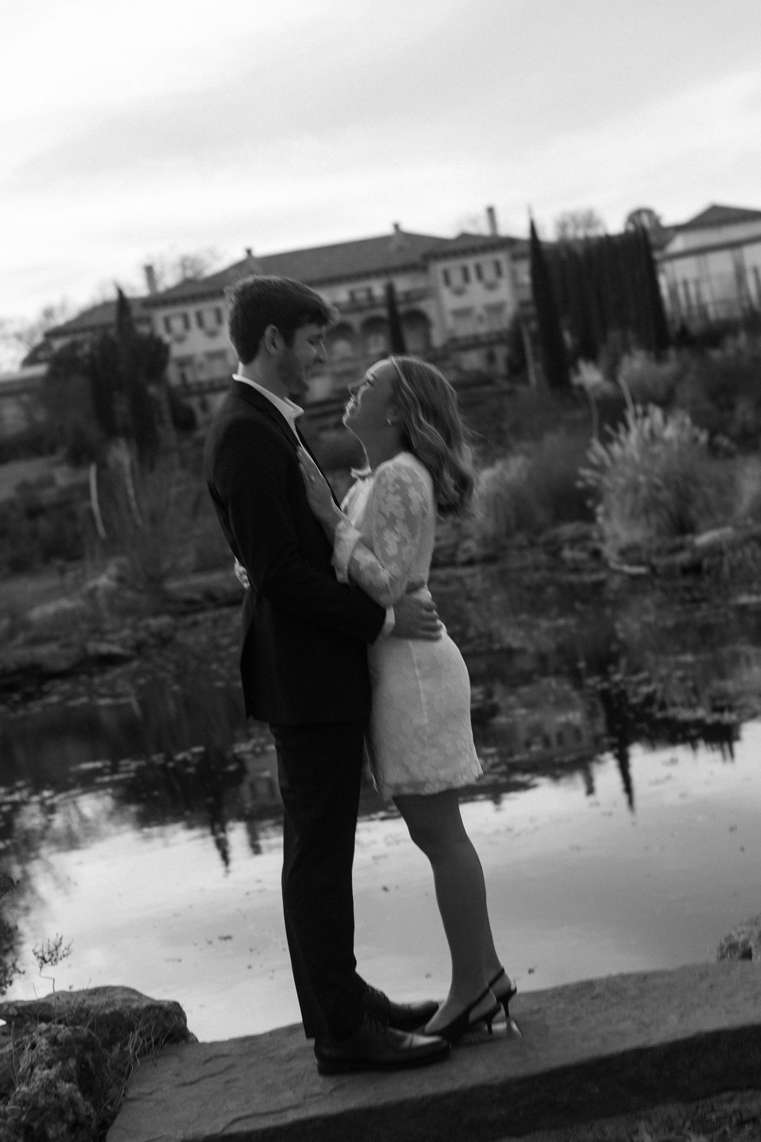 Black and white photos of a couple embracing near water with city buildings in background.