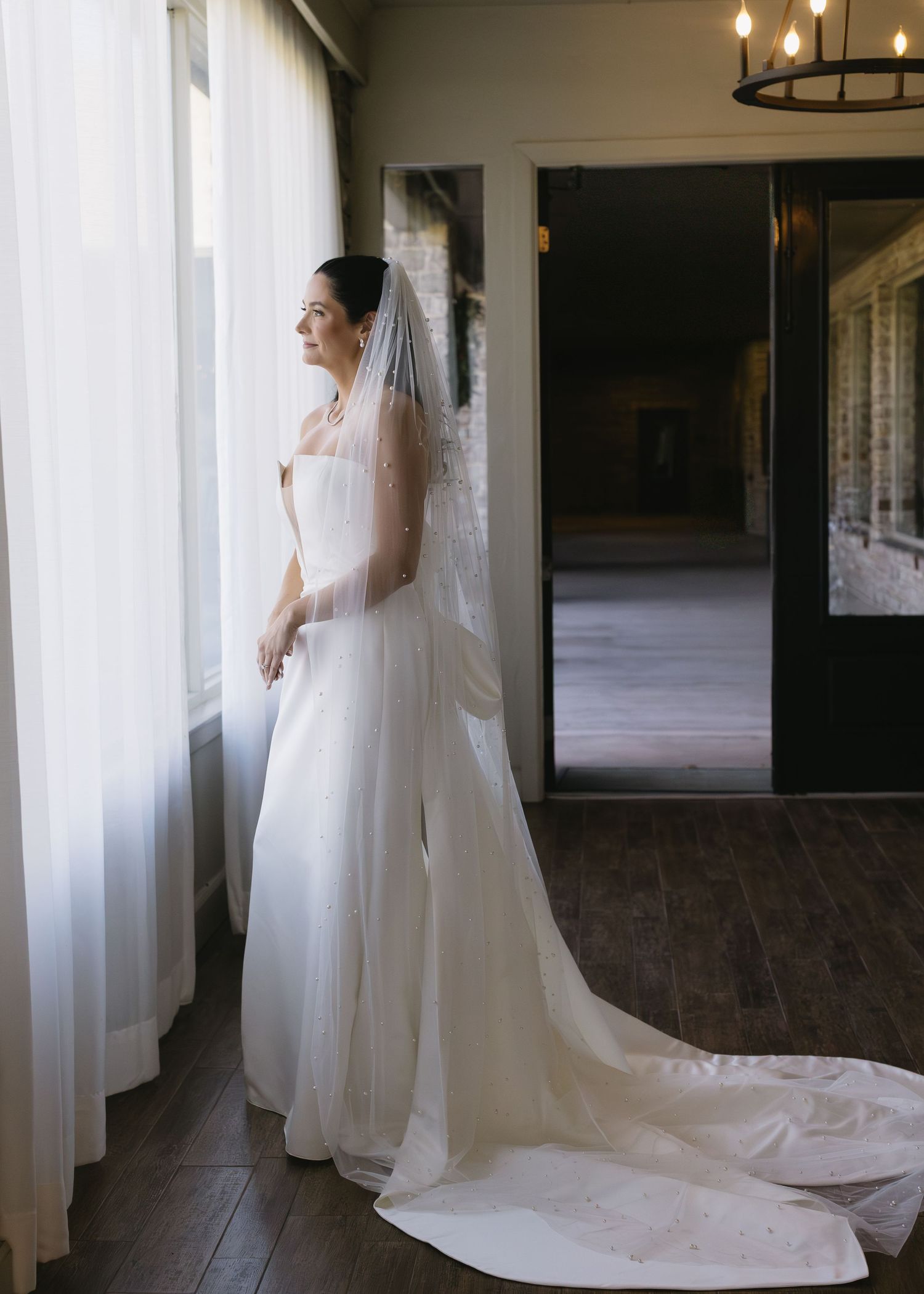 Wedding dress with long train and veil photographed in natural light against white curtains.