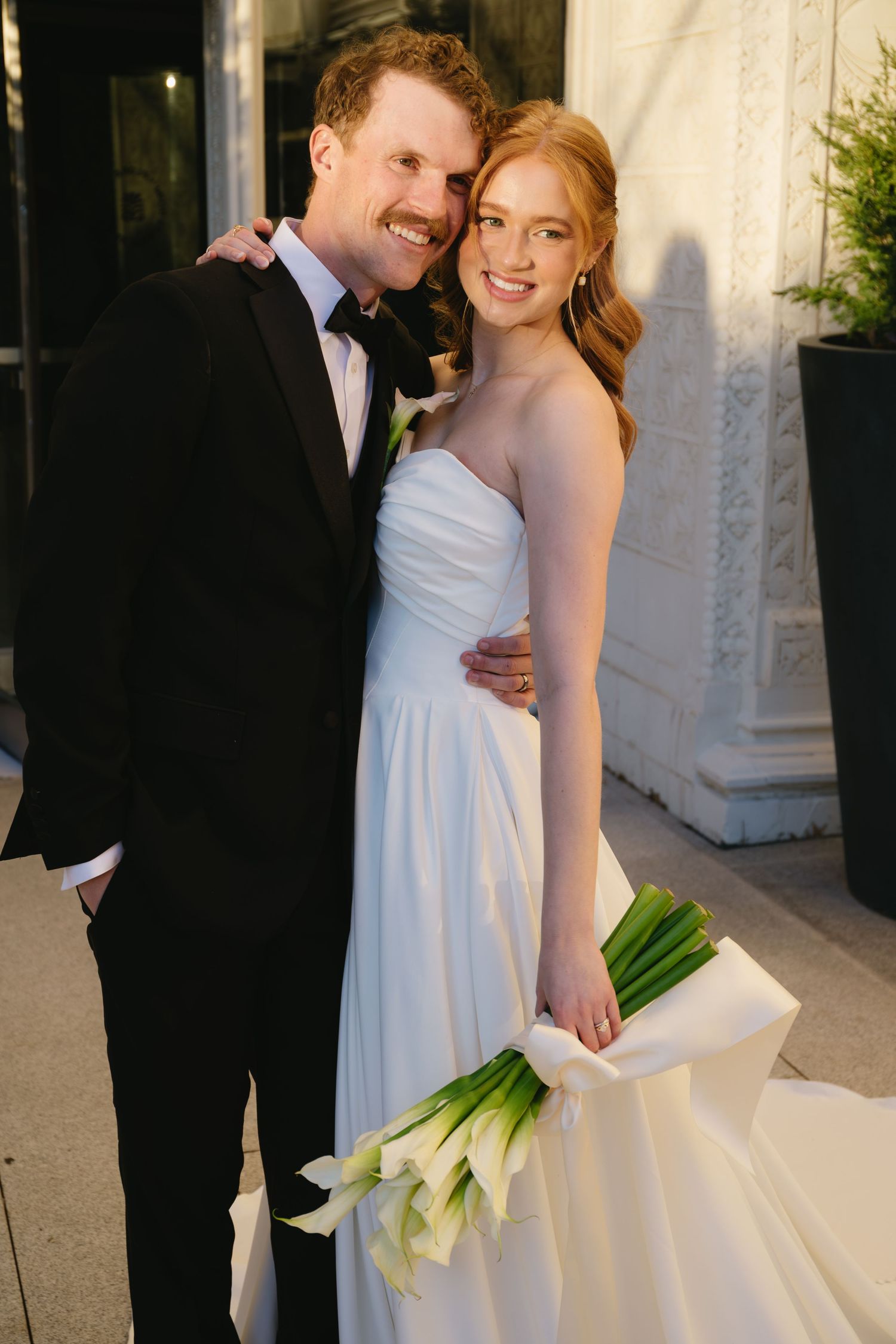 Elegant wedding couple with white calla lily bouquet in outdoor setting.