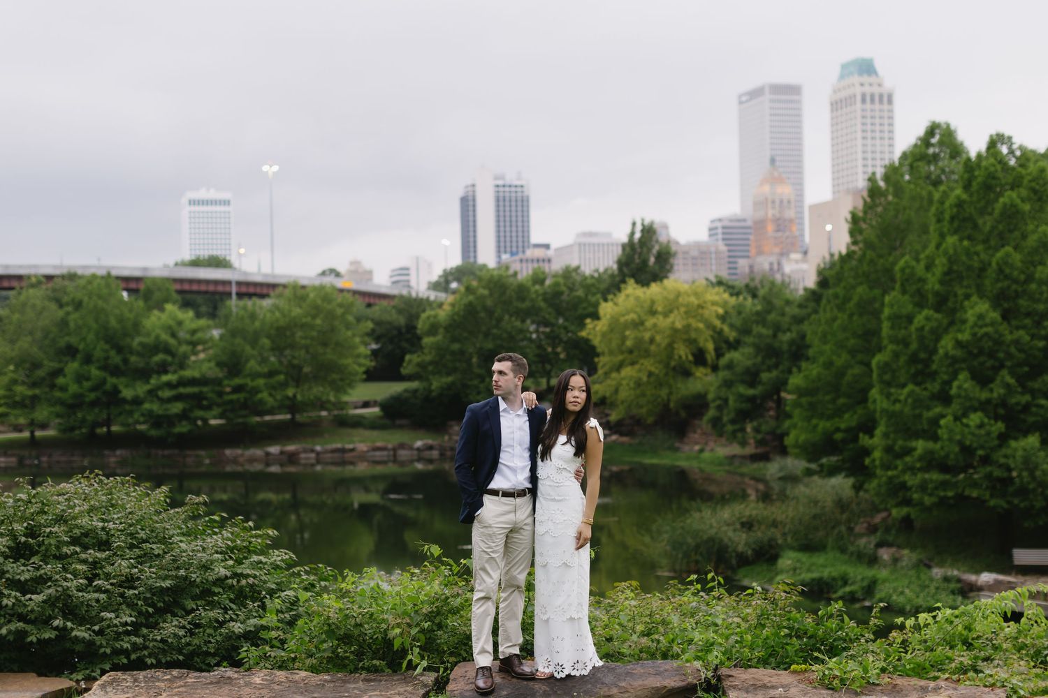 Wedding couple poses against urban skyline backdrop with green park setting.
