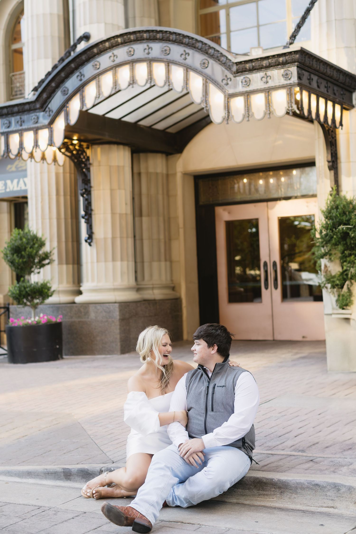 A romantic couple sits on steps outside an elegant building entrance with ornate architectural details and lighting.
