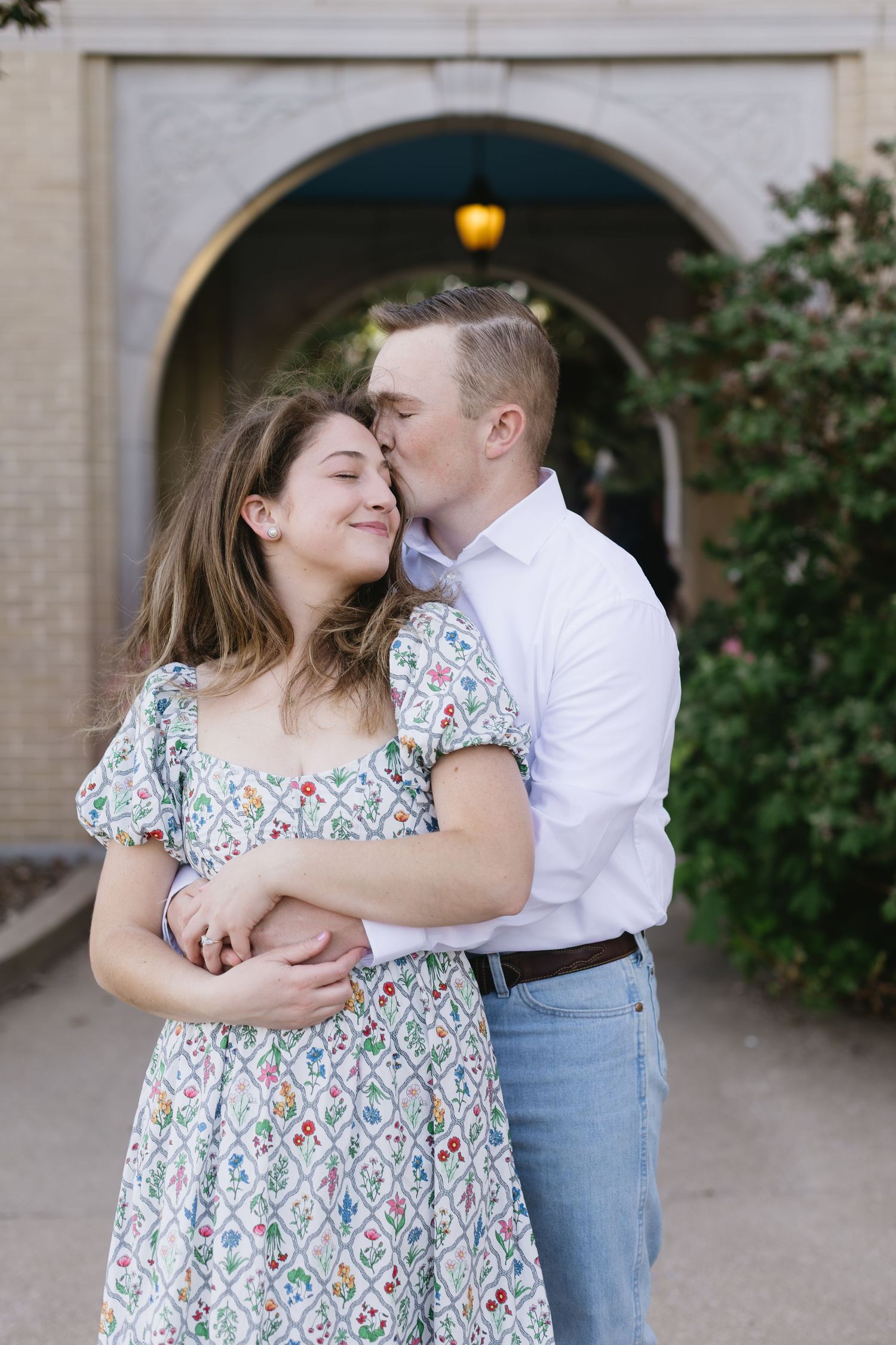 A couple shares tender moments while posing in floral dress and white shirt against an architectural backdrop.