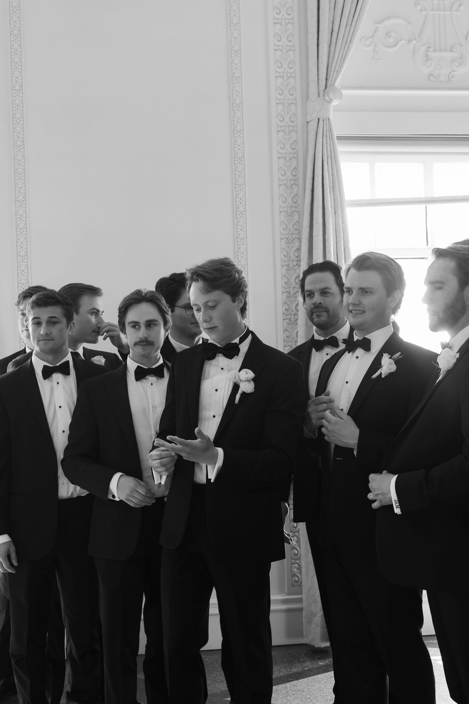 Row of groomsmen in black tuxedos standing in an elegant white room.