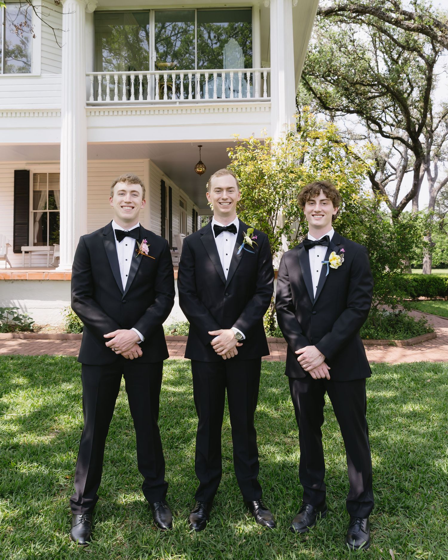 Three groomsmen in black tuxedos and bow ties stand on a lawn in front of a white columned porch.