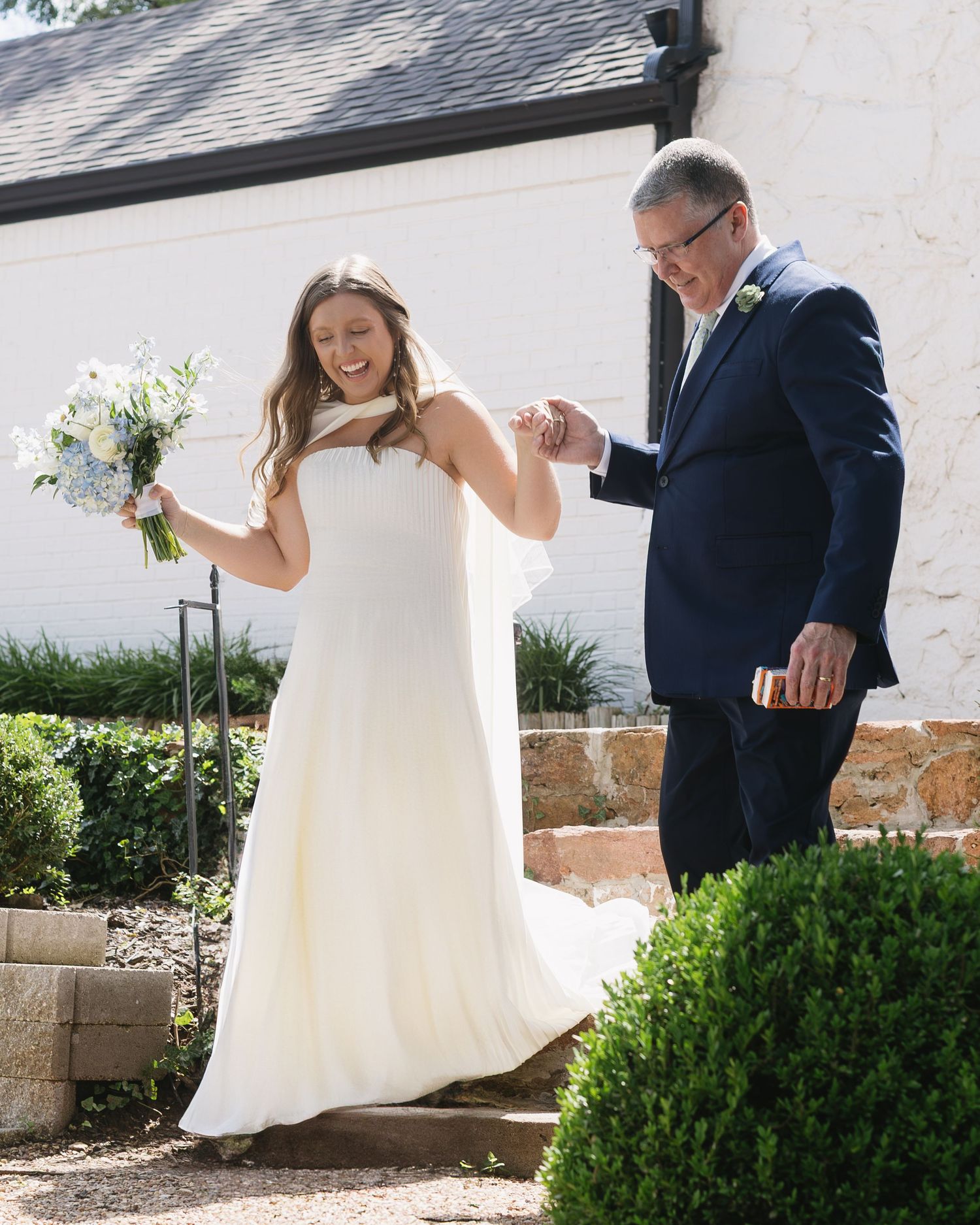 Wedding couple playfully dances on a stone patio with white building and greenery in background.