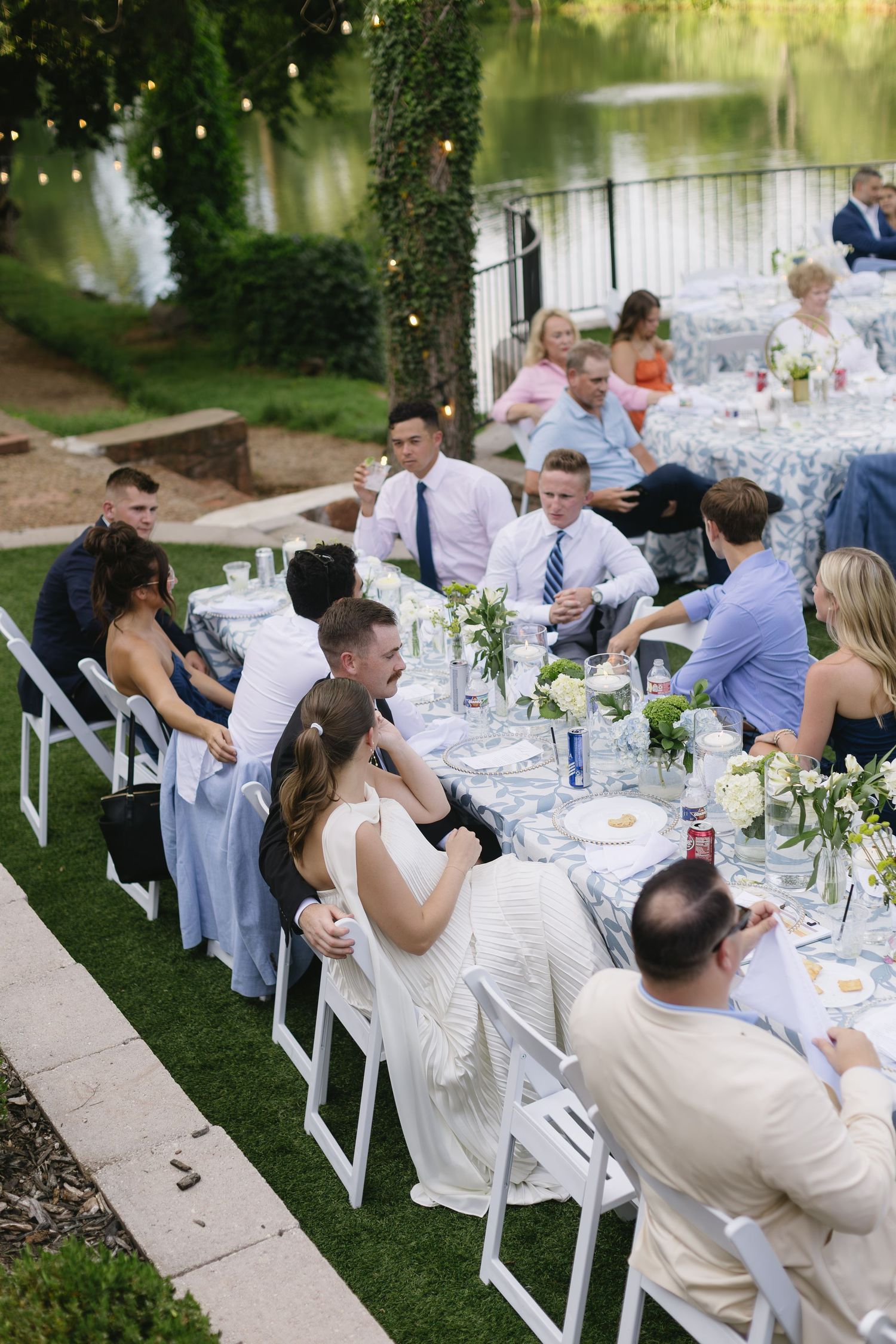 Outdoor wedding reception with guests seated at long white tables under trees beside a lake.