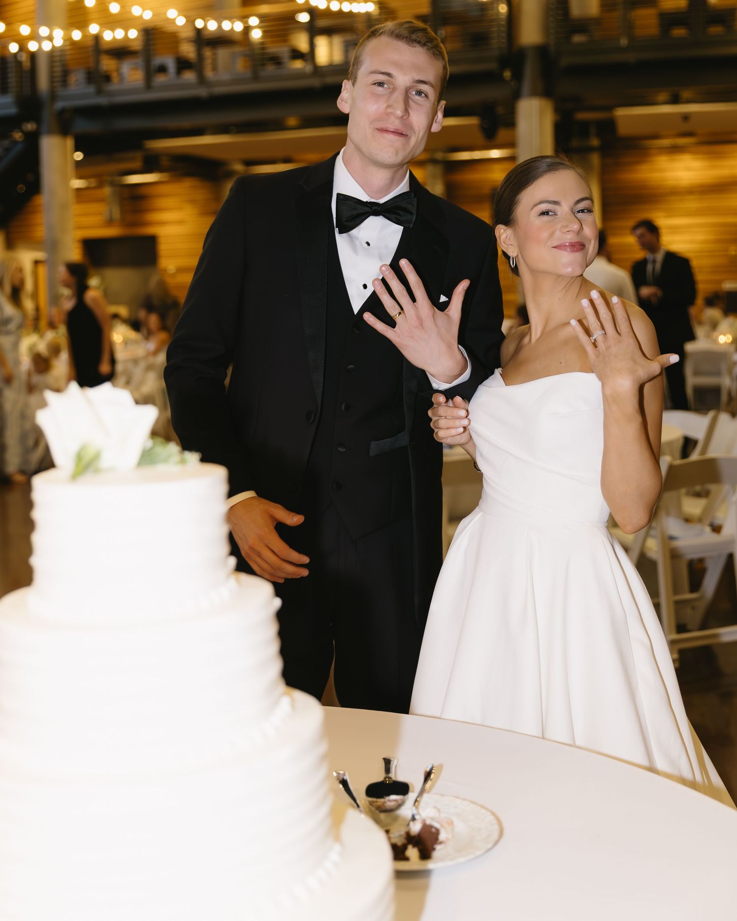 A wedding couple shows off their rings next to a white tiered cake at their reception.
