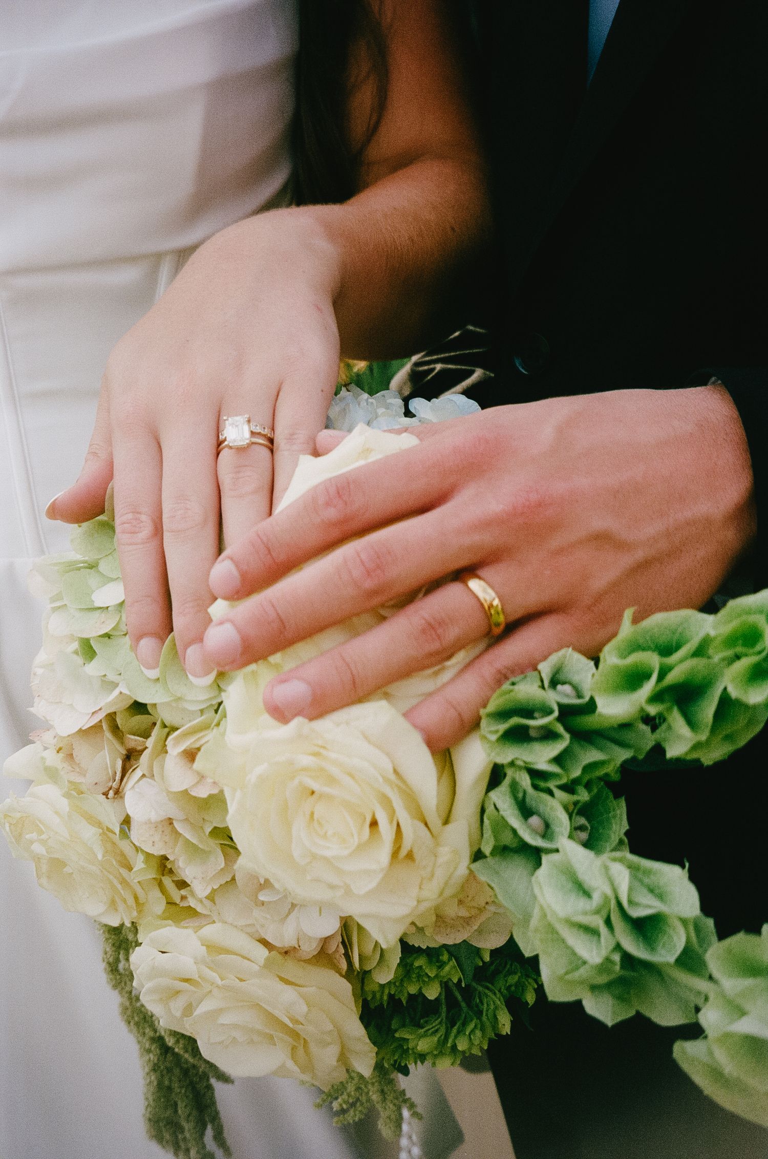 Close-up of hands holding a white and green wedding bouquet with rings visible.