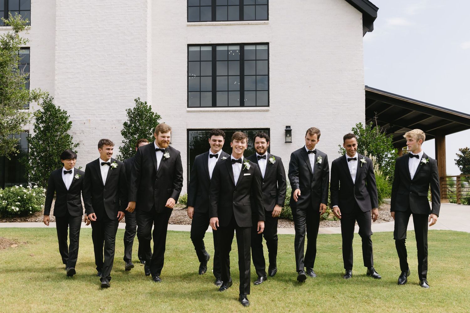 Groomsmen in black tuxedos pose playfully on grass outside modern white building.