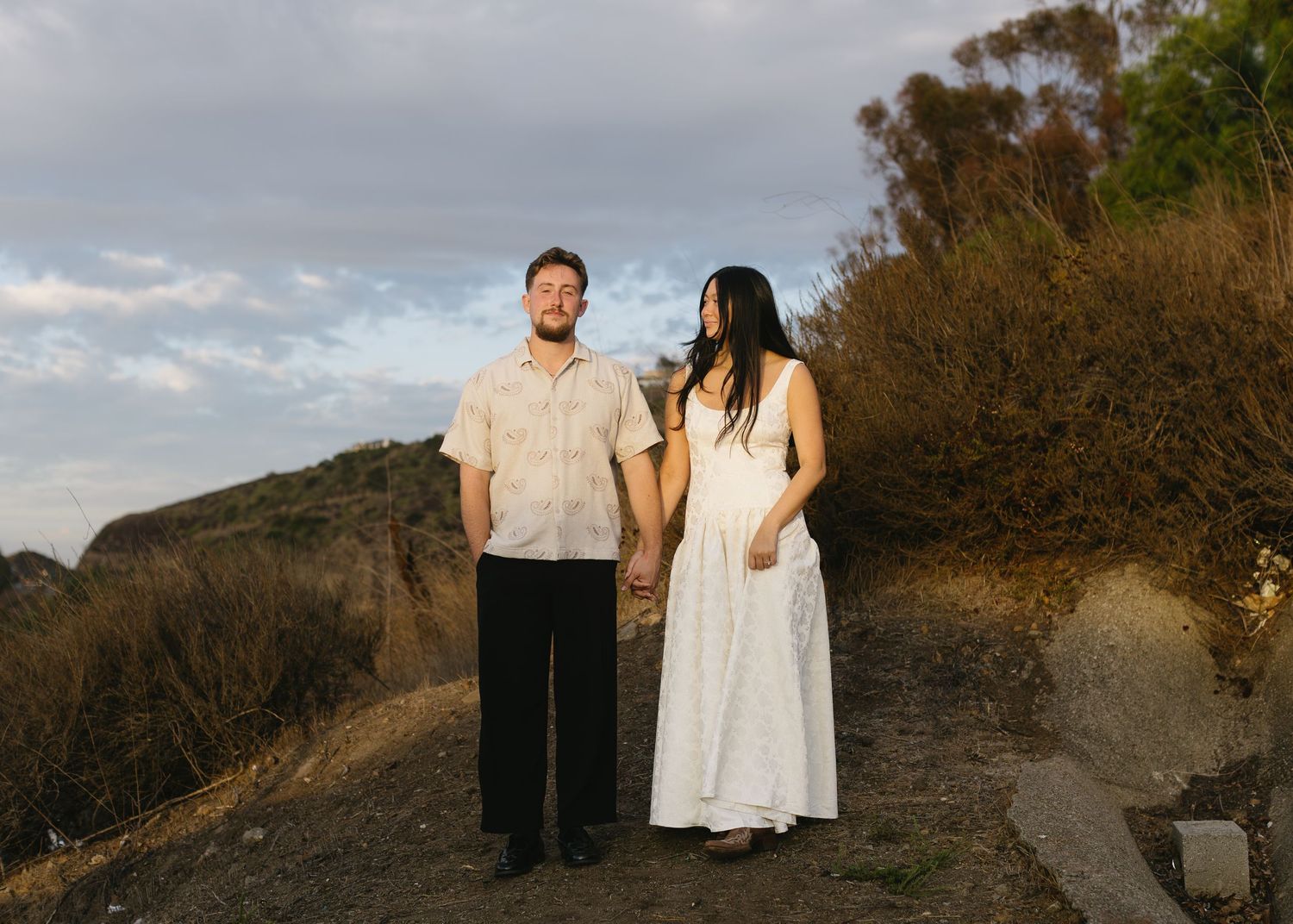A couple in light-colored clothing stands together on a hillside trail during golden hour.