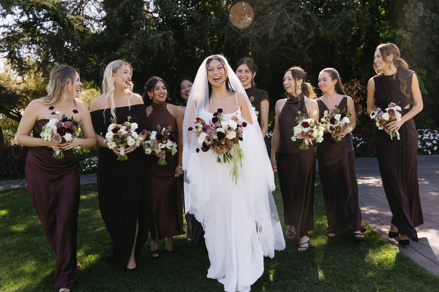 A bridal party in dark brown dresses poses outdoors with white and purple floral bouquets.
