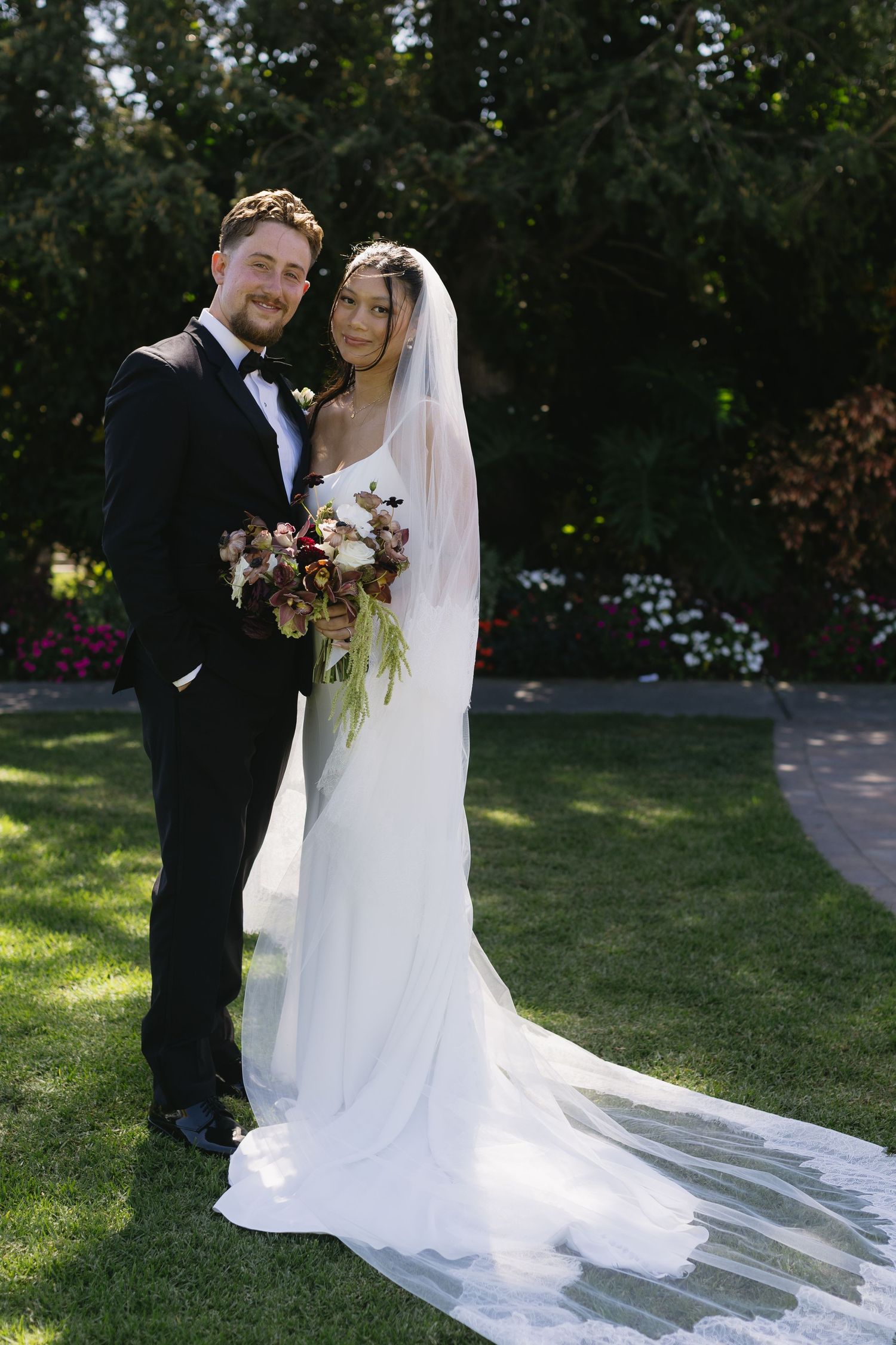 Wedding couple poses together on sunny lawn with flowing white veil and bouquet.