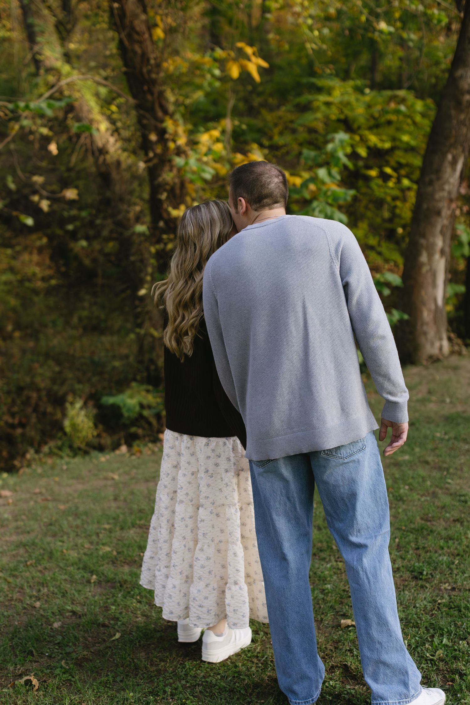 A couple walking away hand in hand on a grassy slope with fall foliage in the background.