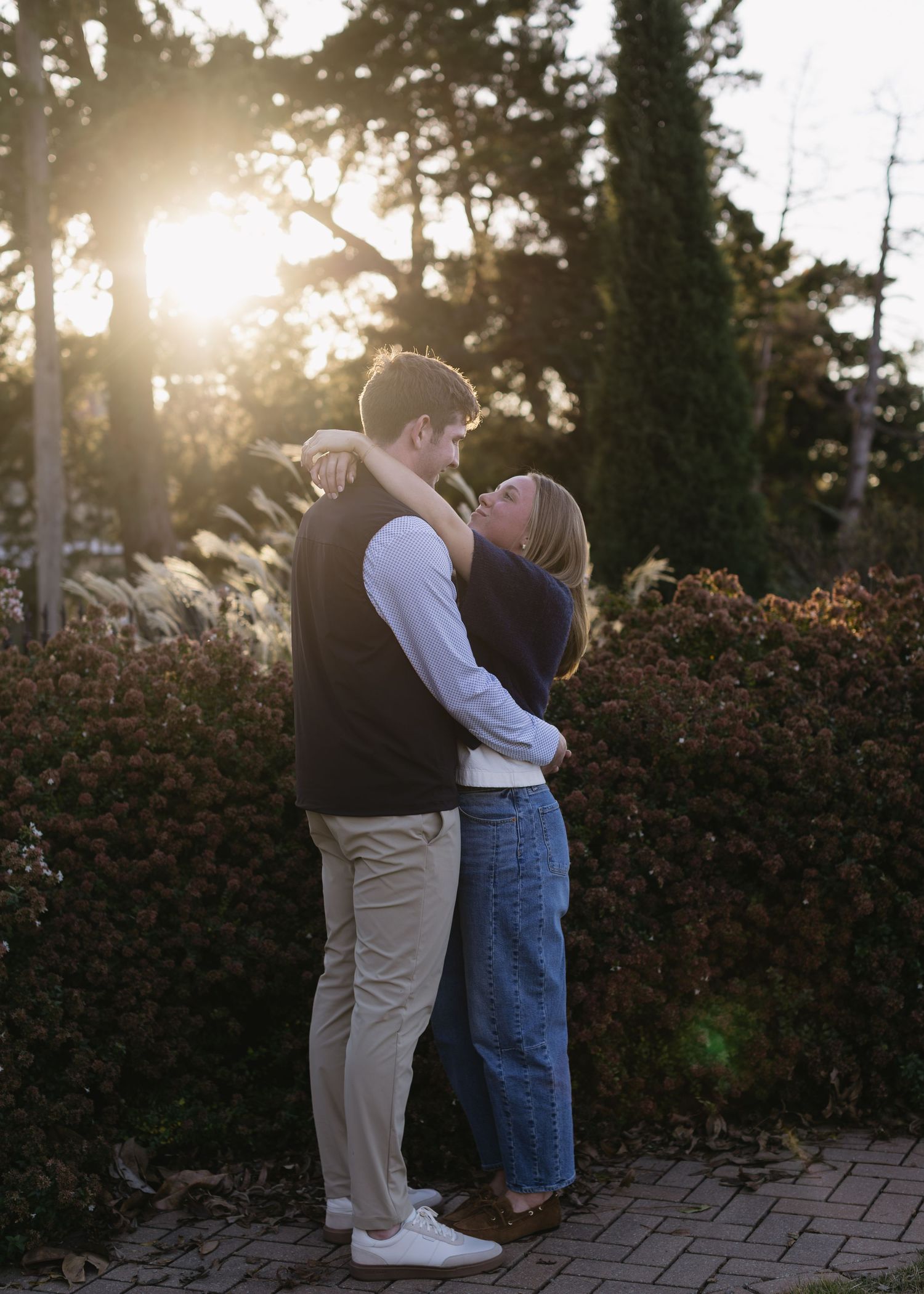 Two people share an intimate moment against a backdrop of hedges and sunlight.