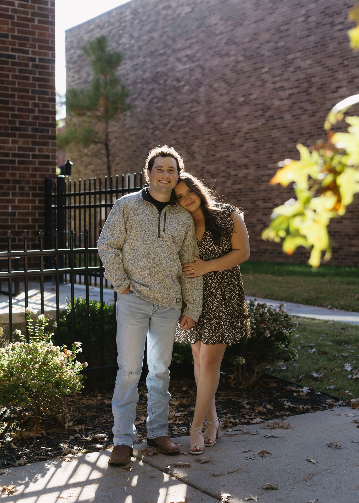 A couple embraces in casual wear while standing in front of a brick wall on a sunny day.