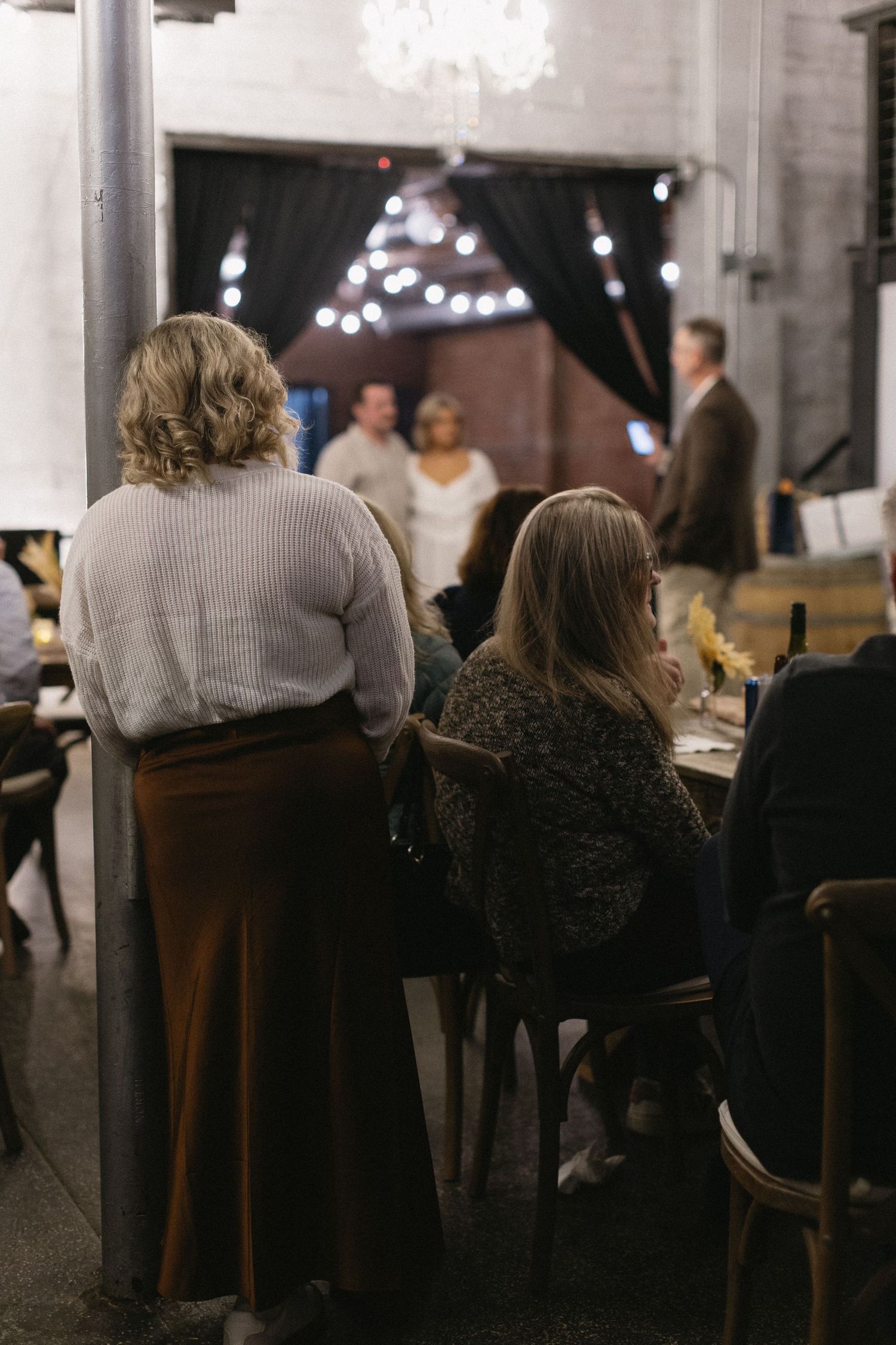 Wedding guests seated at tables watching ceremony in industrial venue with string lights.