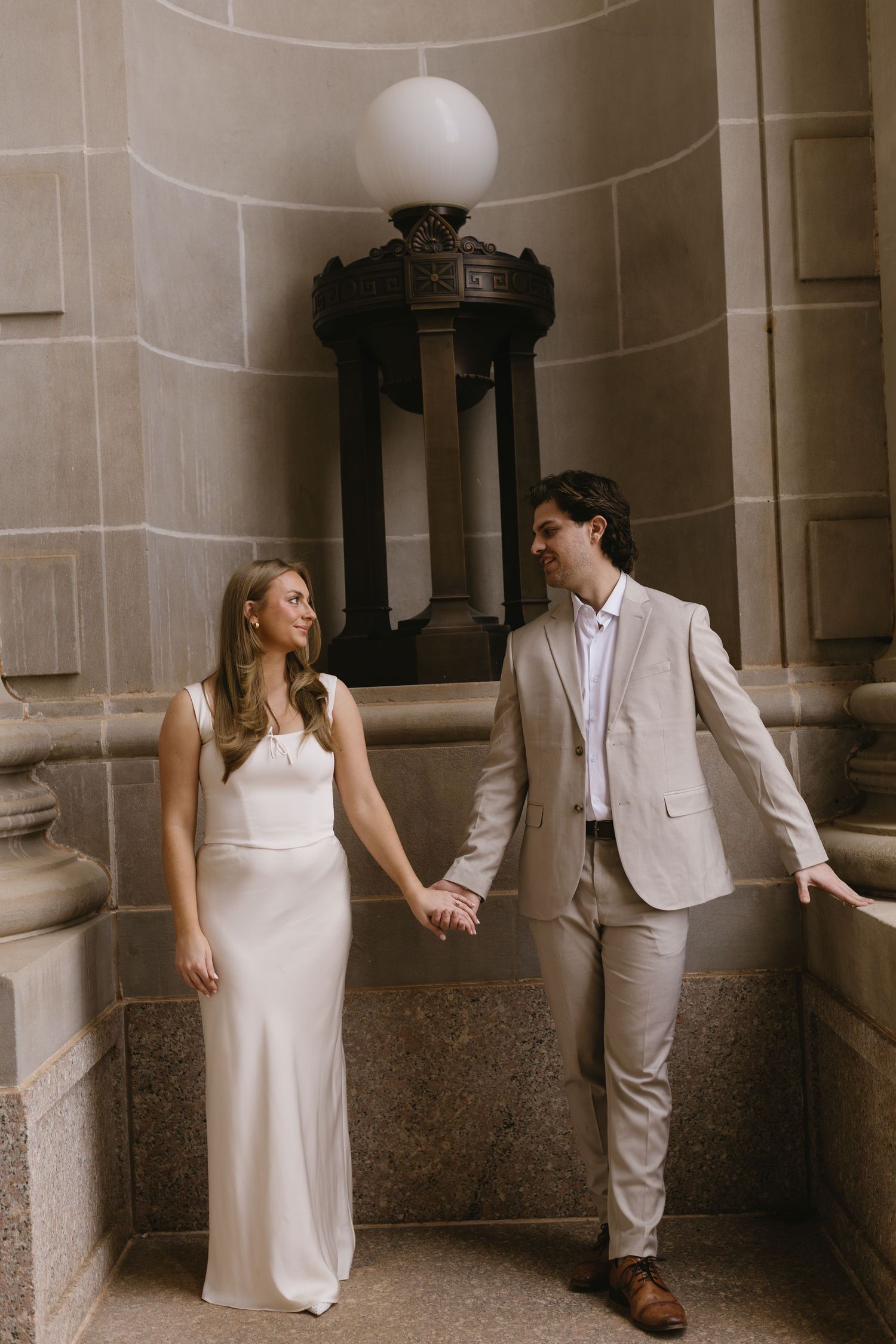 A couple in coordinated formal wear poses playfully in an elegant hallway with columns.