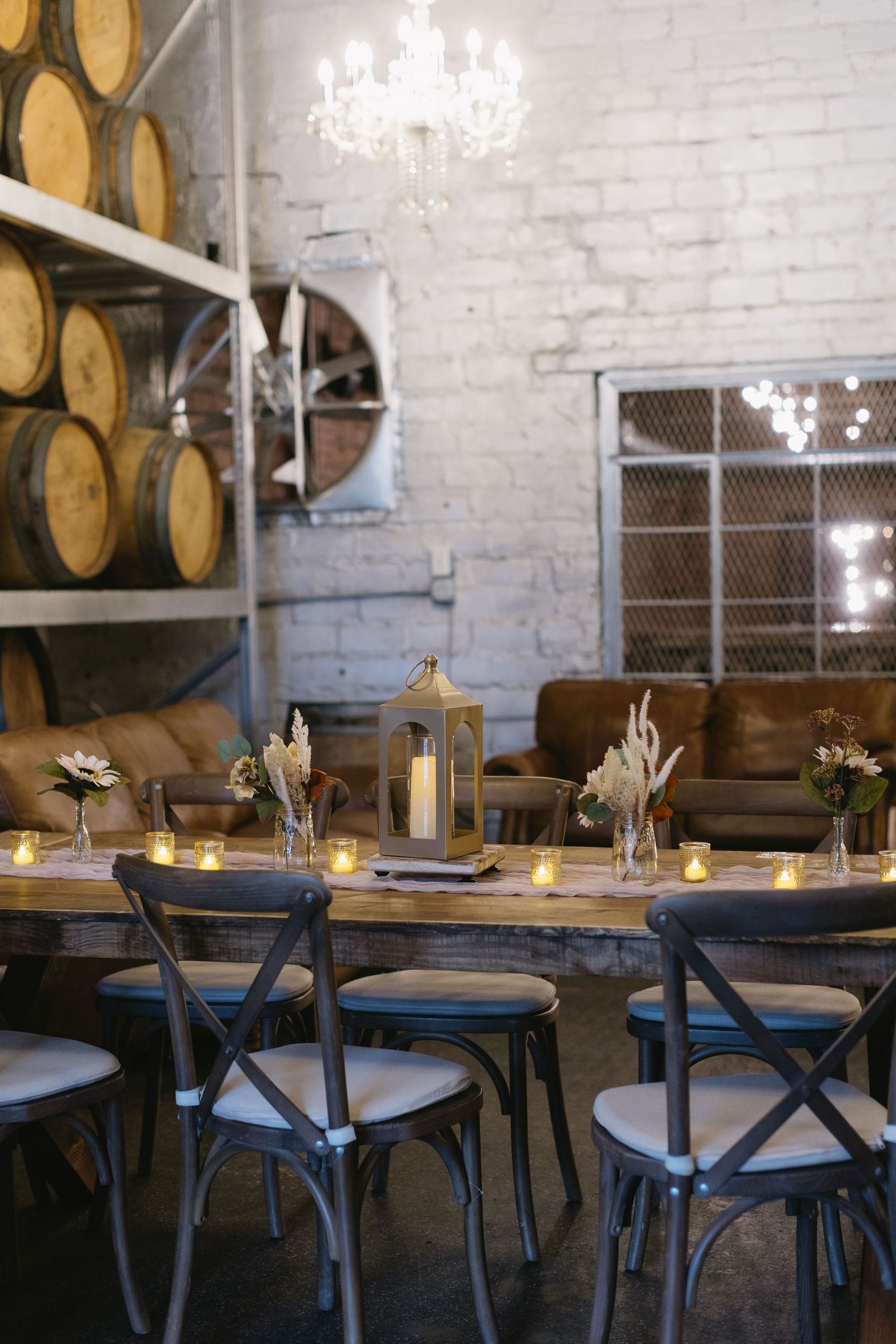 Rustic dining area with wooden barrels, crystal chandeliers, and industrial chairs in a wine cellar setting.