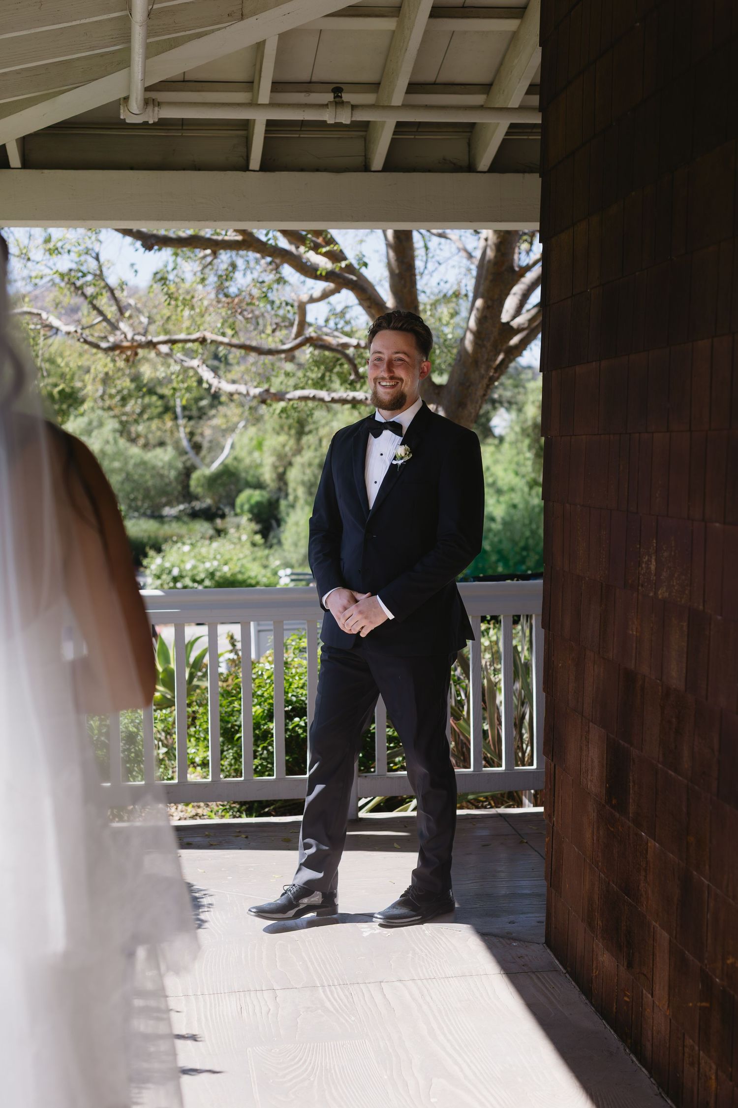 Groom in classic black tuxedo and bowtie stands on wooden porch.