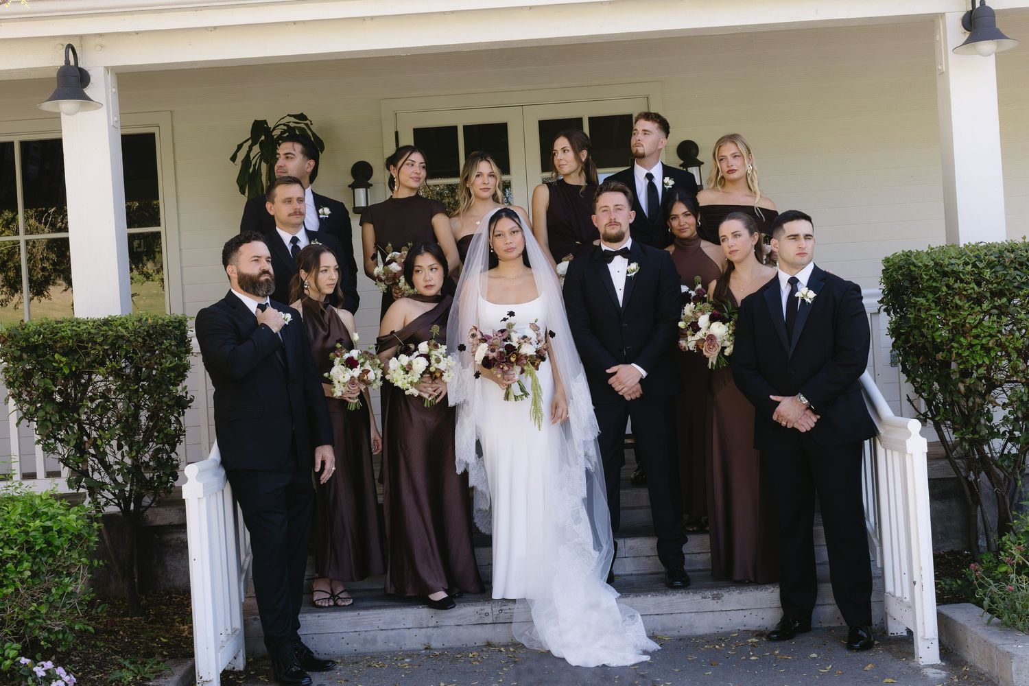 Large wedding party poses on white porch for formal group photo.