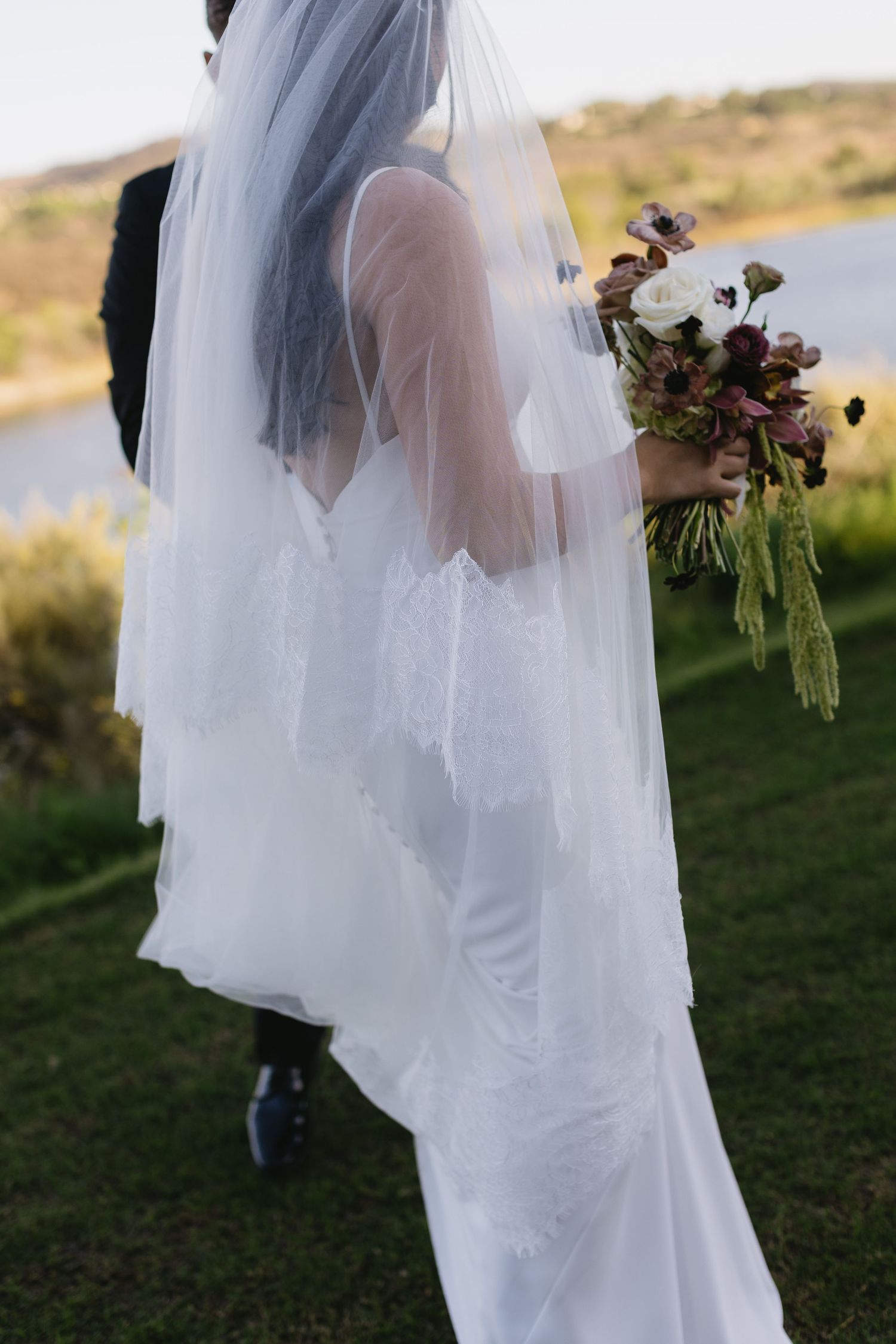 Bride in flowing white veil holds bouquet against scenic outdoor backdrop.