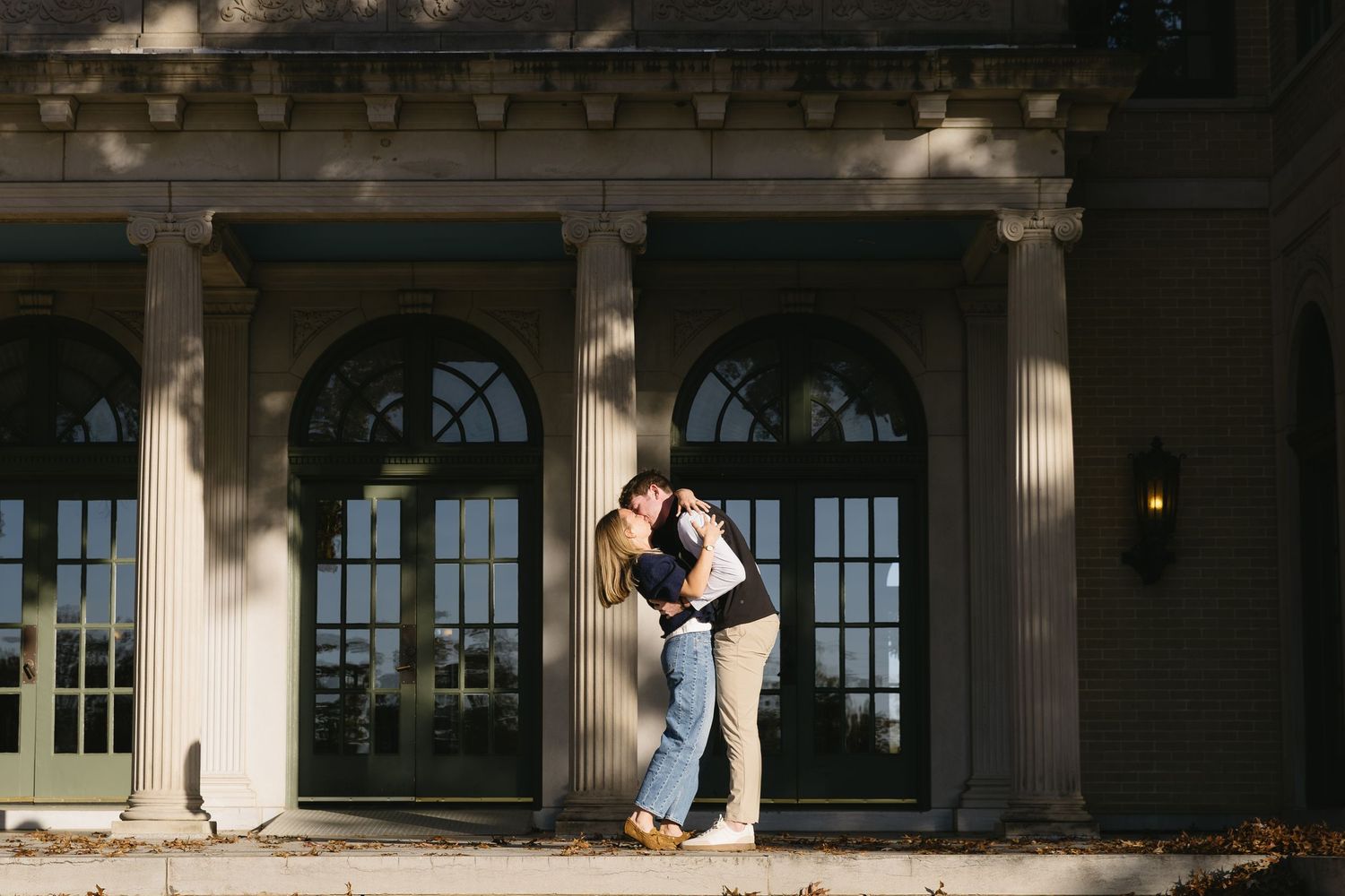 A romantic couple embraces on a porch with elegant columns and arched windows in the background.