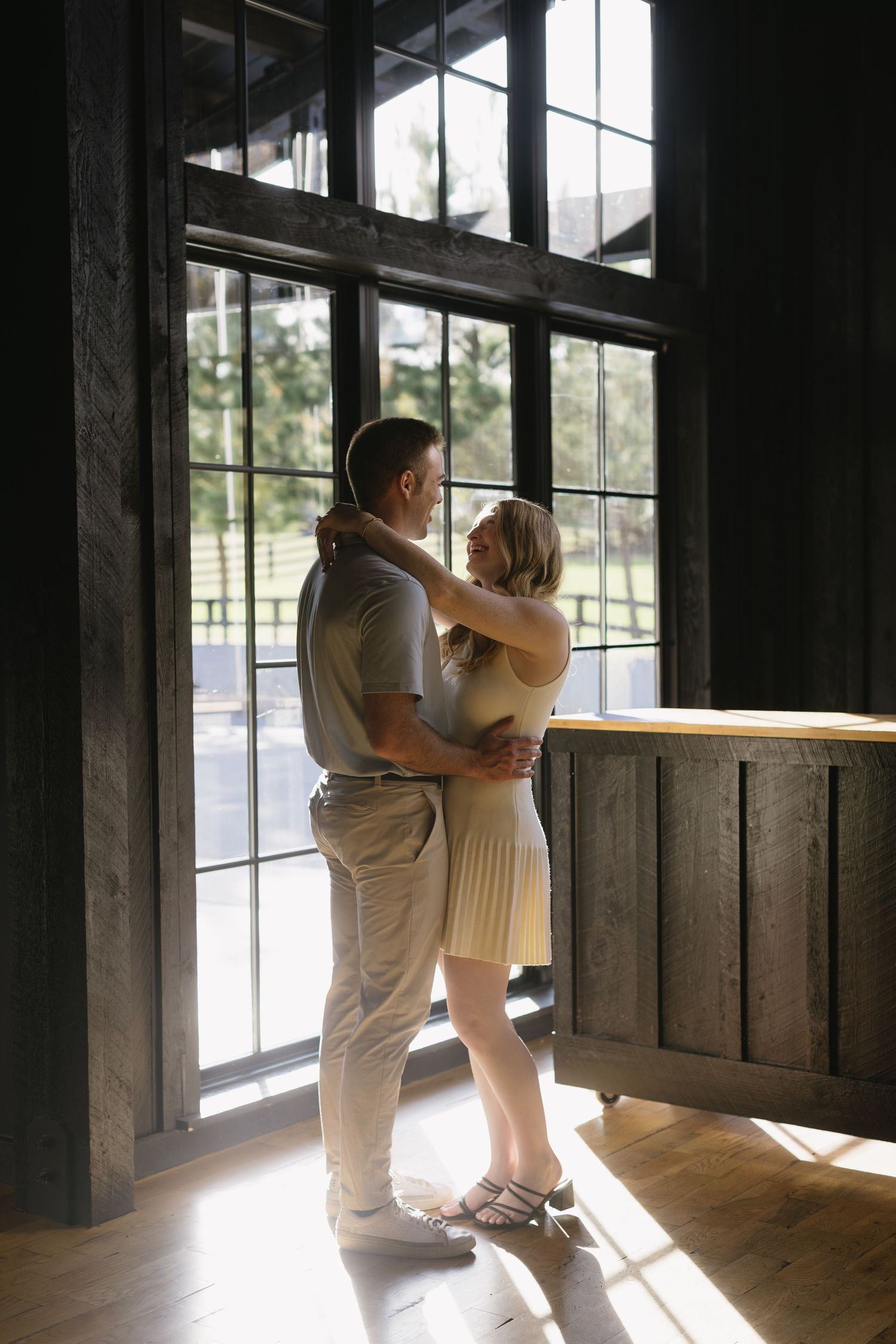A romantic couple in white clothing embraces in a rustic wooden barn with natural light streaming through windows.