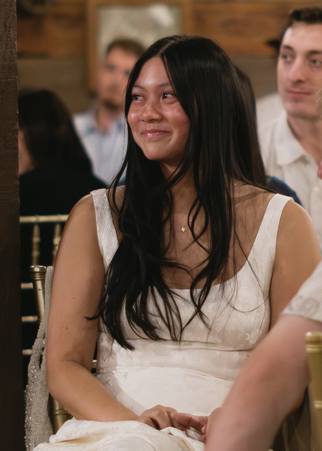A person in a white sleeveless top smiles while sitting at what appears to be a dining or event setting.