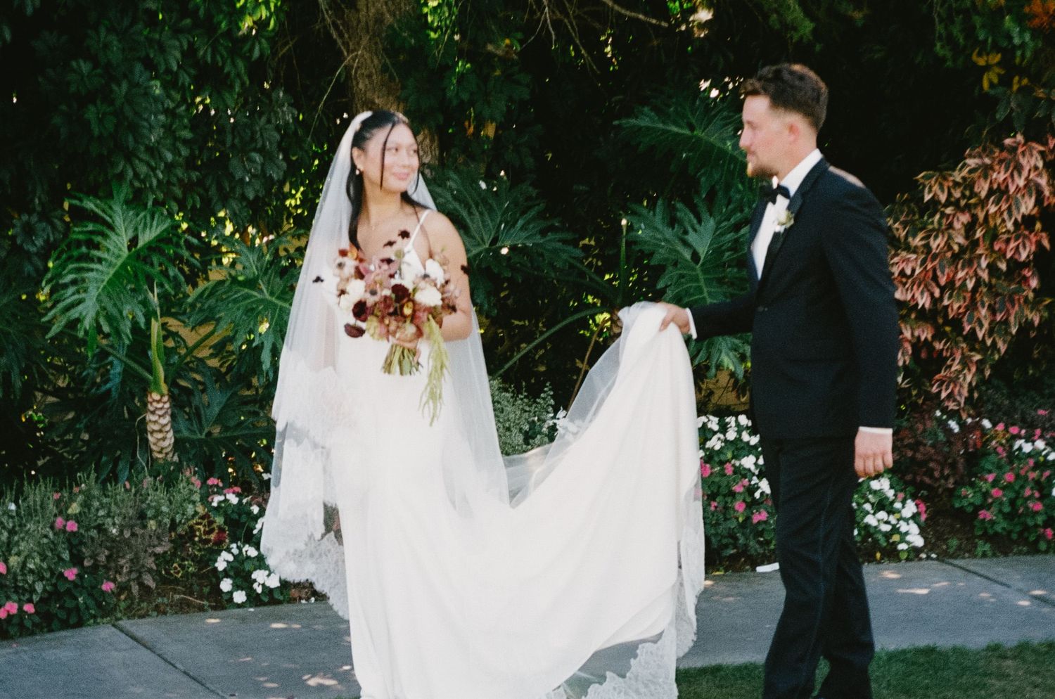 Bride and groom walk together through lush garden setting during outdoor wedding ceremony.