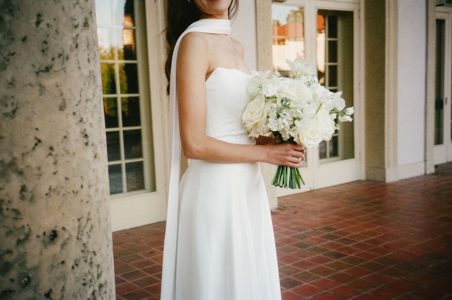 A bride in a white wedding dress holds a bouquet while standing on a brick porch with white columns.