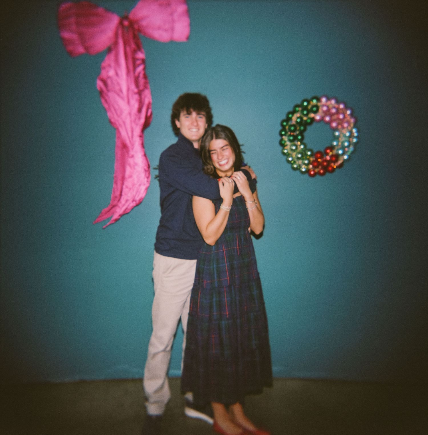 A couple dancing playfully with pink fabric flowing through the air against a turquoise blue wall with a decorative wreath.
